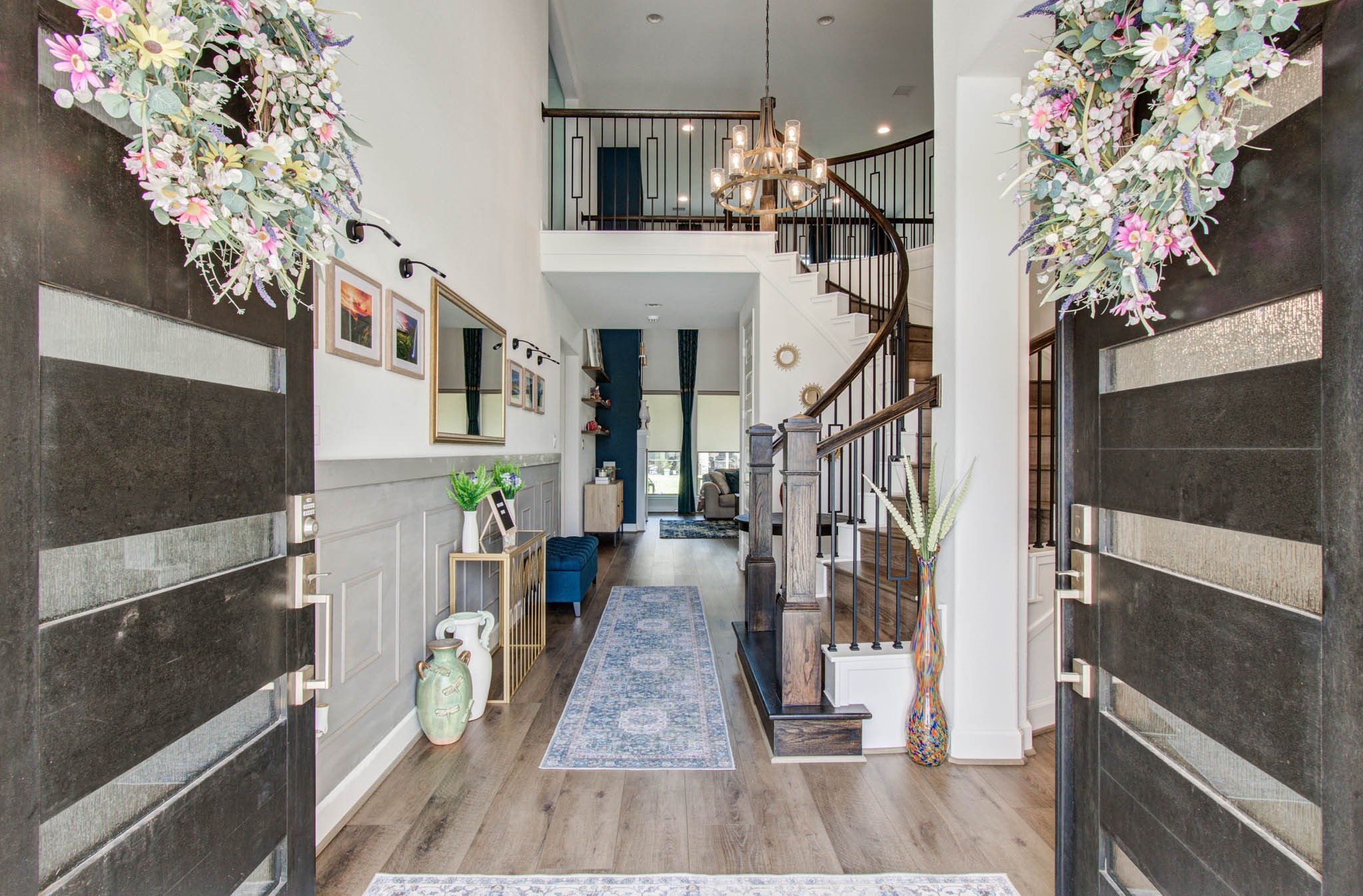 Step into elegance through grand double doors with floral wreaths, revealing a bright foyer with wood flooring and a graceful curved staircase, all enhanced by stylish light fixtures and decorative touches for a sophisticated ambiance.