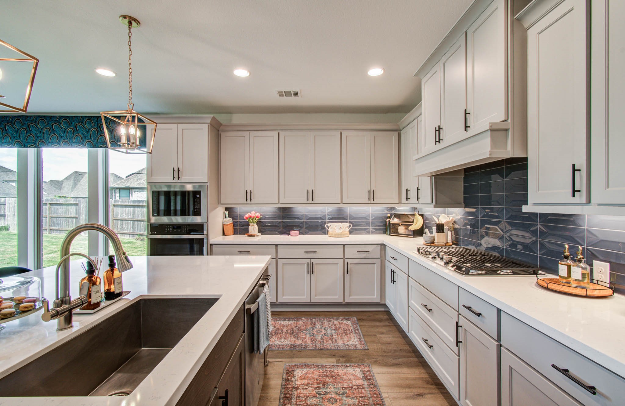 A modern kitchen with sleek white cabinetry, a spacious island featuring a sink, and stainless steel appliances is beautifully illuminated by large windows and stylish lighting, creating an inviting and functional space perfect for cooking and entertaining.
