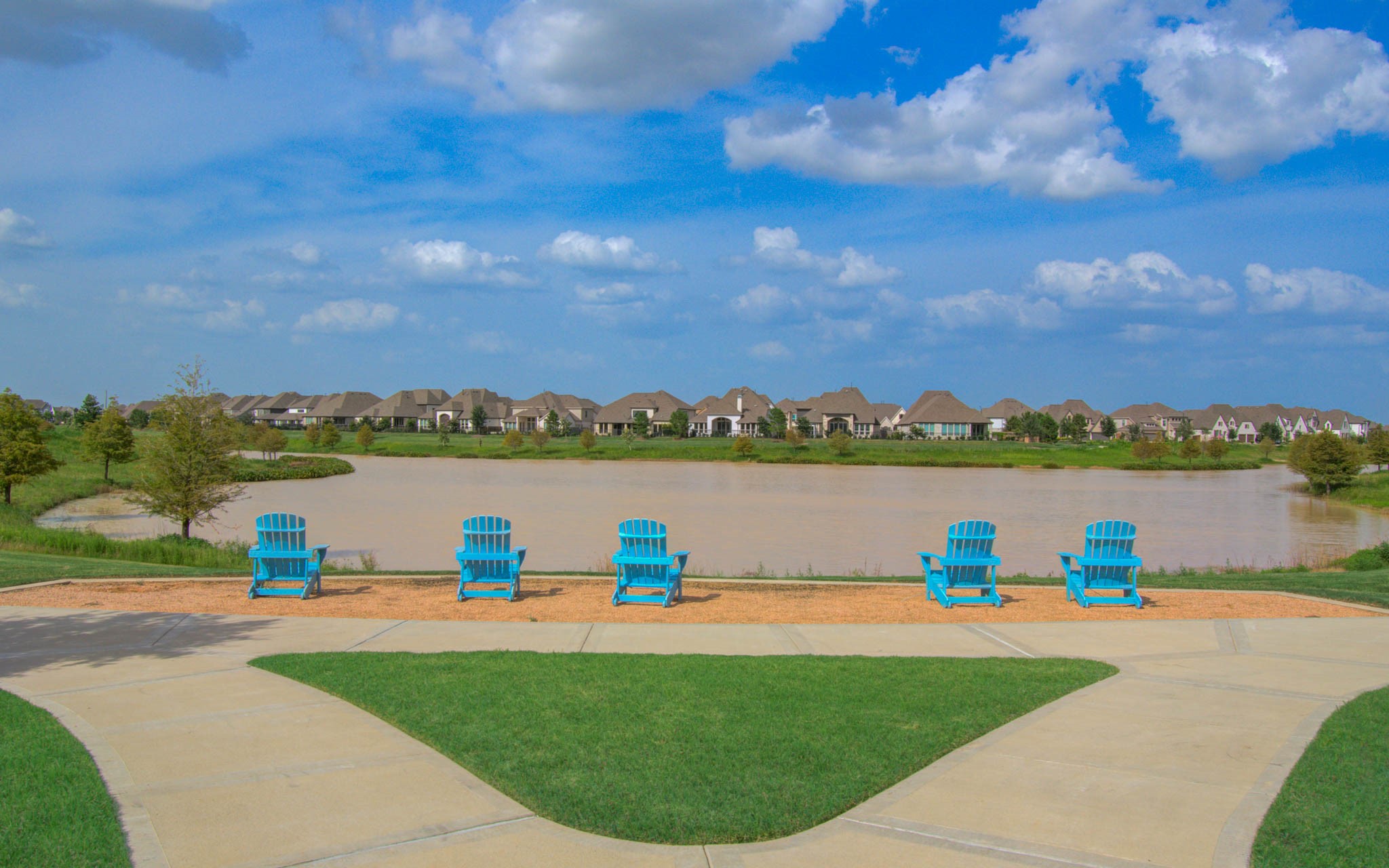 Experience ultimate relaxation with this serene lakeside view, featuring a row of inviting blue Adirondack chairs perfectly positioned to overlook the calm water and picturesque neighborhood homes under a bright blue sky.