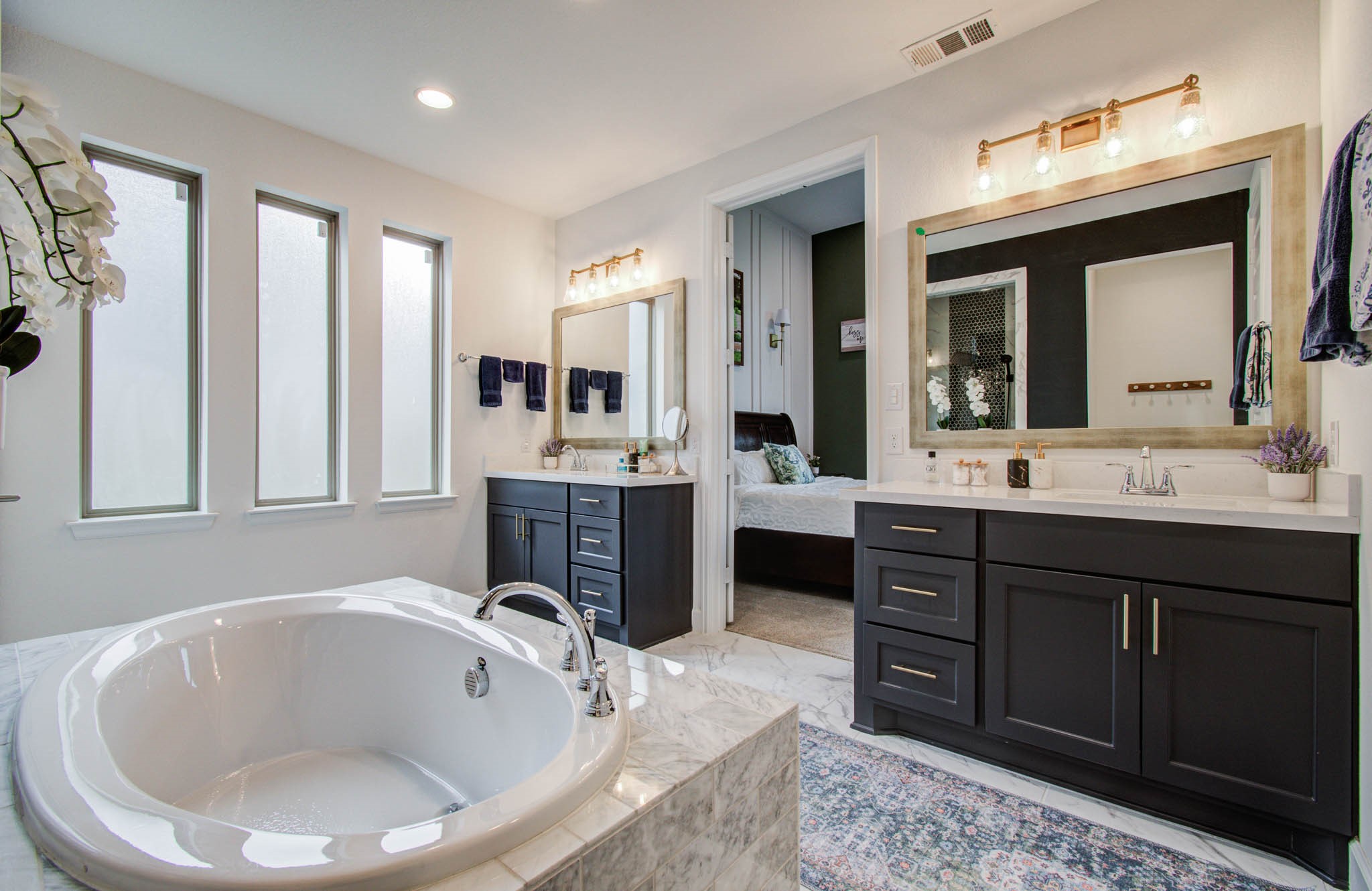 This elegant bathroom features a spacious bathtub, dual vanities with dark cabinetry, and large windows for natural light. It offers a modern design with stylish fixtures, leading into a connected bedroom for convenience.