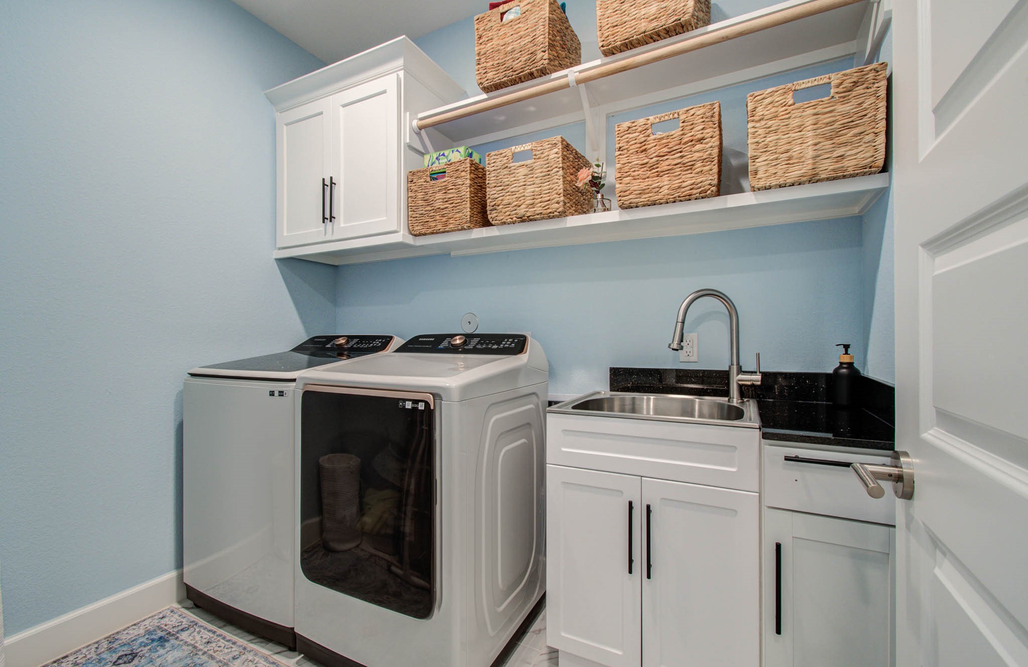 This bright and functional laundry room features modern appliances, ample storage with overhead cabinets and woven baskets, a convenient sink, and a sleek black countertop, all set against a soft blue wall.