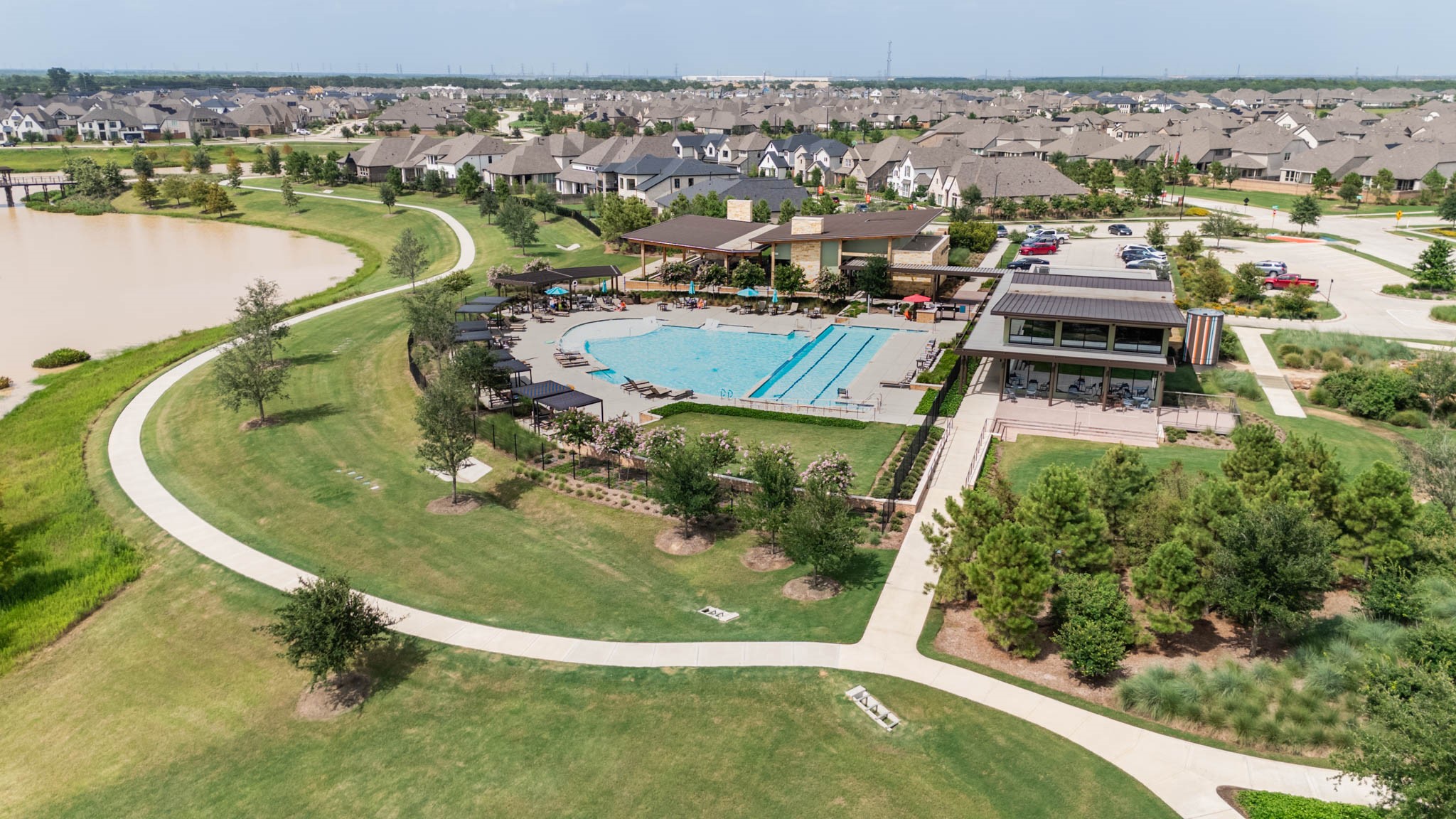 This photo showcases a vibrant community with a spacious outdoor recreational area featuring a large swimming pool, green spaces, and a scenic pond. The neighborhood is composed of modern, well-maintained homes, offering a tranquil yet active lifestyle.