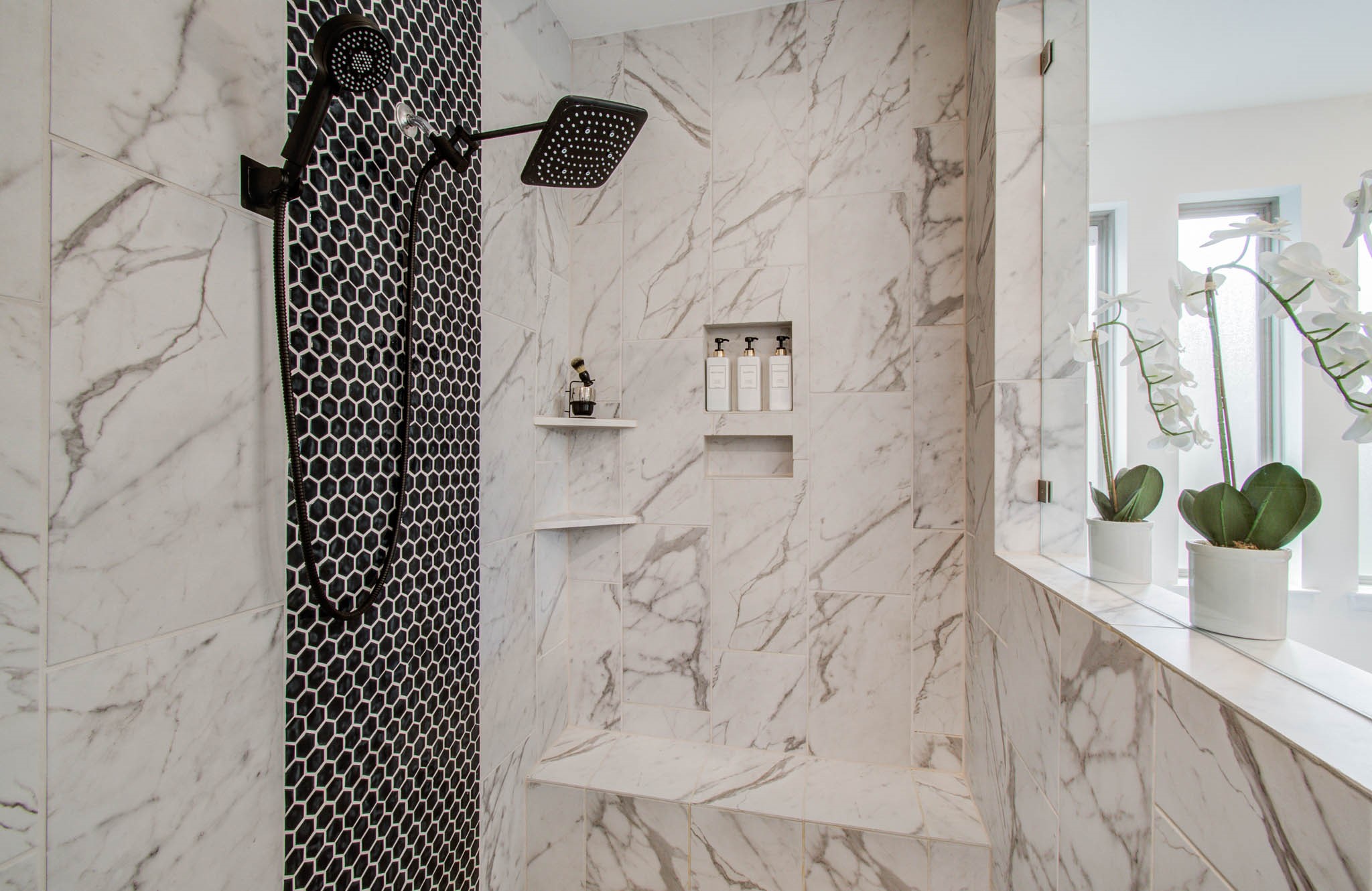 Experience ultimate luxury in this bright, airy shower featuring elegant marble tiles, a striking black hexagonal accent wall, a modern rain showerhead, and convenient built-in shelving and bench.