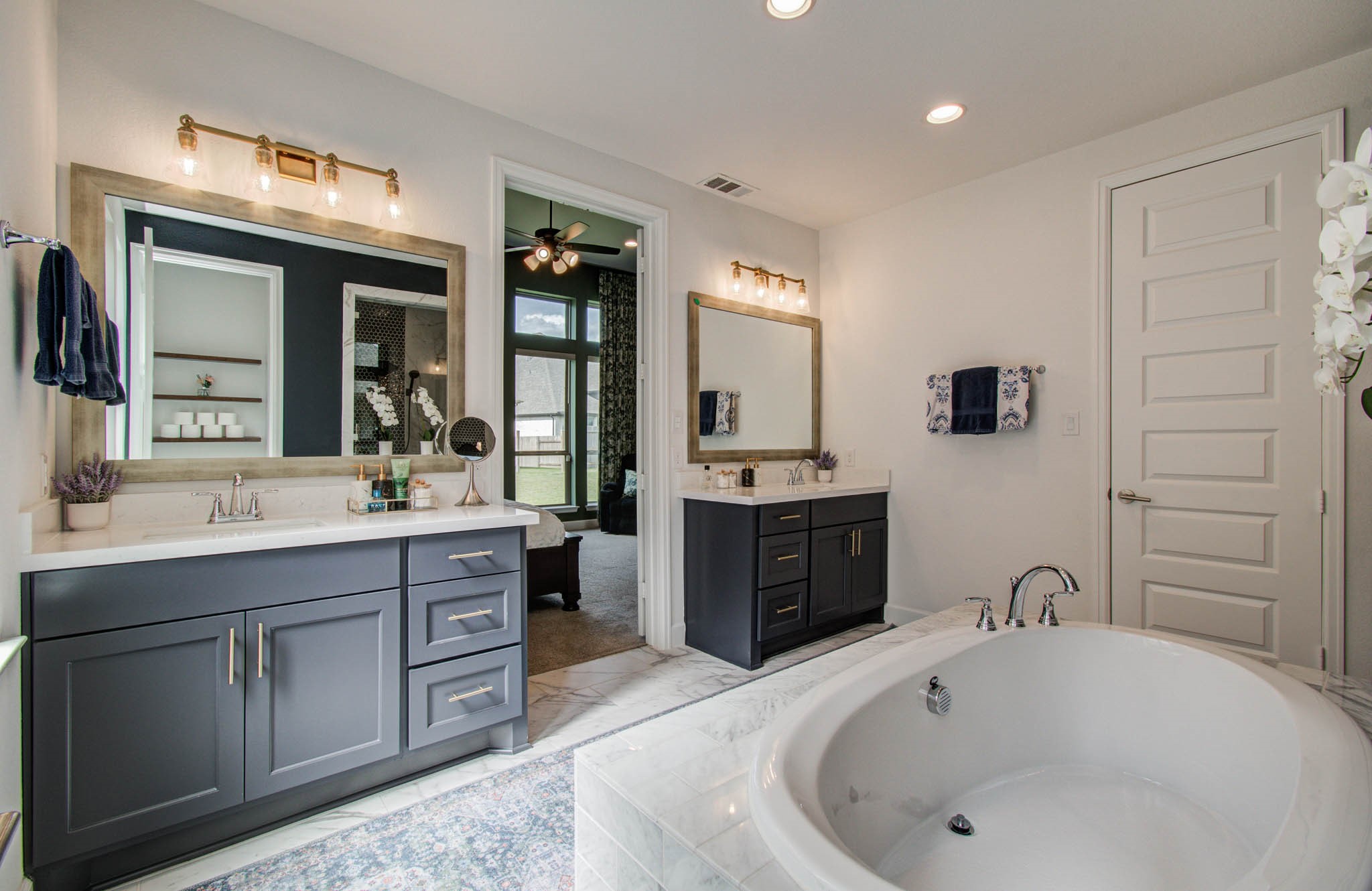This spacious bathroom features dual vanities with modern fixtures, a large soaking tub, and elegant lighting. Neutral tones and a glimpse into an adjoining room create a serene, cohesive atmosphere.