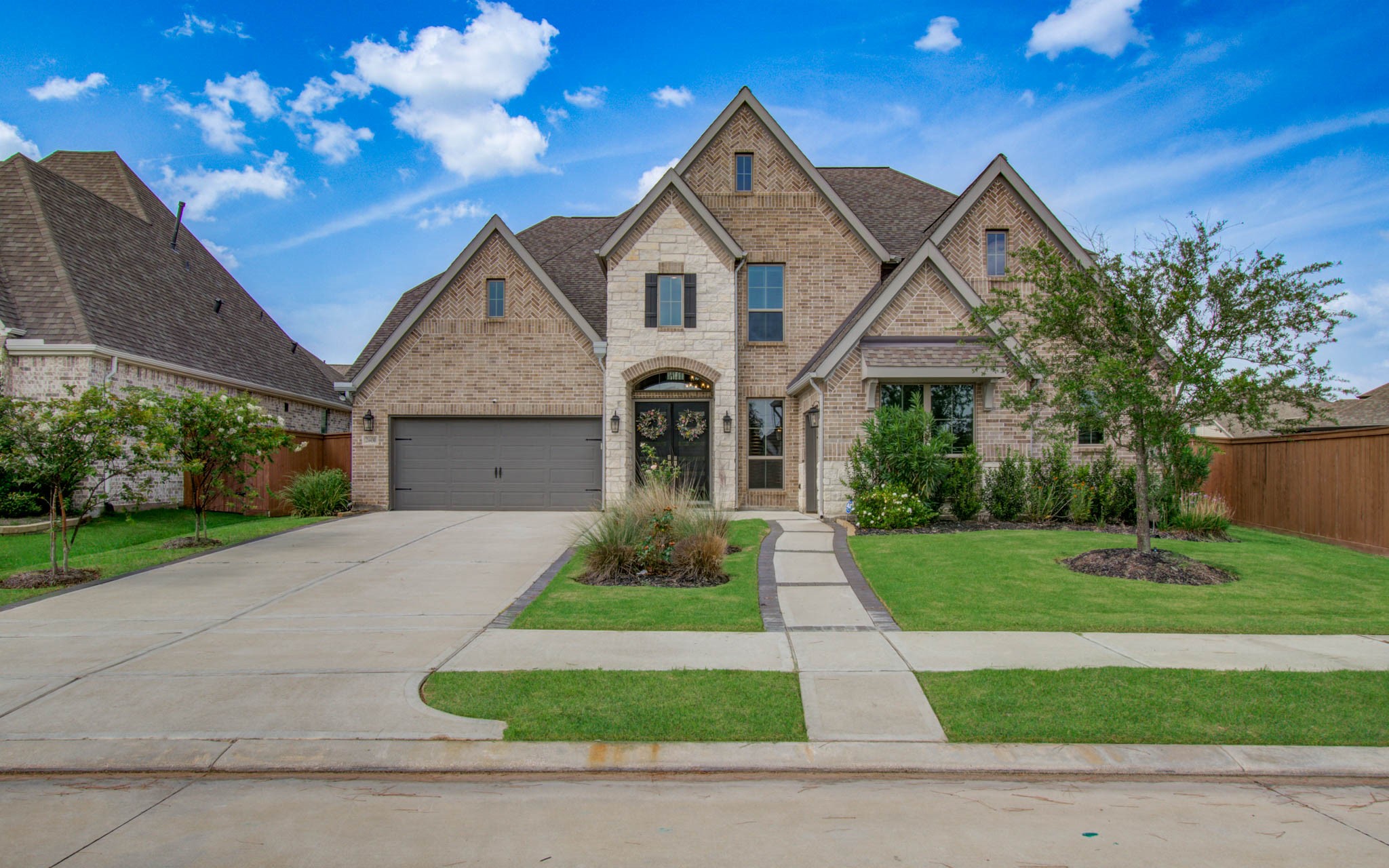 This is a beautiful two-story brick home with a steeply pitched roof, featuring a spacious driveway leading to a two-car garage. The well-maintained front yard includes a paved walkway, manicured lawn, and mature trees, offering excellent curb appeal.