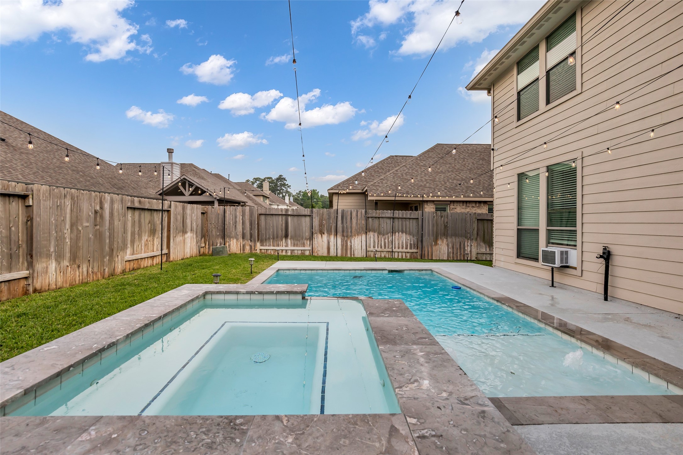 This backyard features a modern pool and spa, surrounded by a wooden fence for privacy. String lights add ambiance, and there's a grassy area for relaxation.