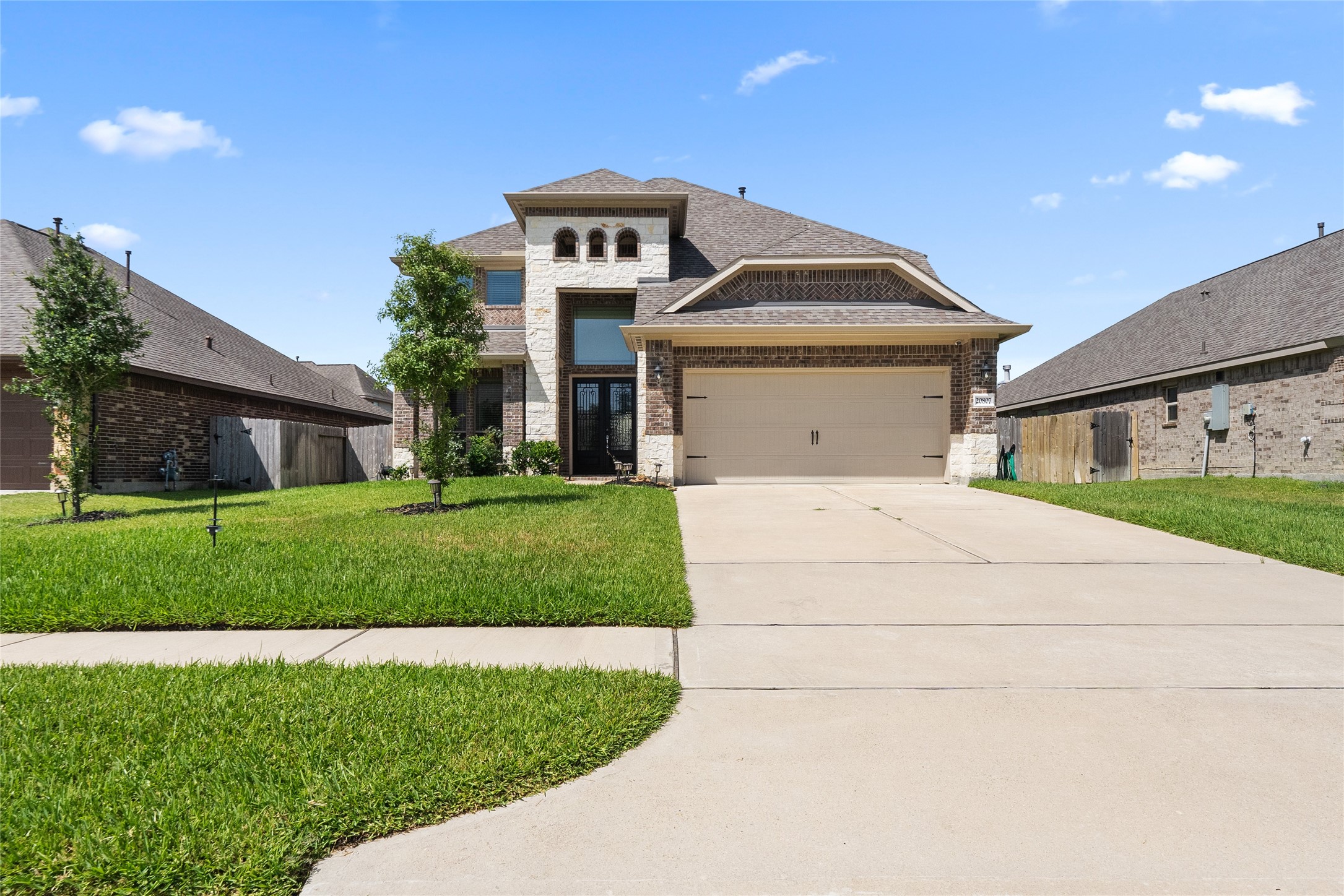 This spacious two-story home features a modern brick and stone facade, a double garage, and a well-maintained front yard with young trees and lush grass, set in a suburban neighborhood.