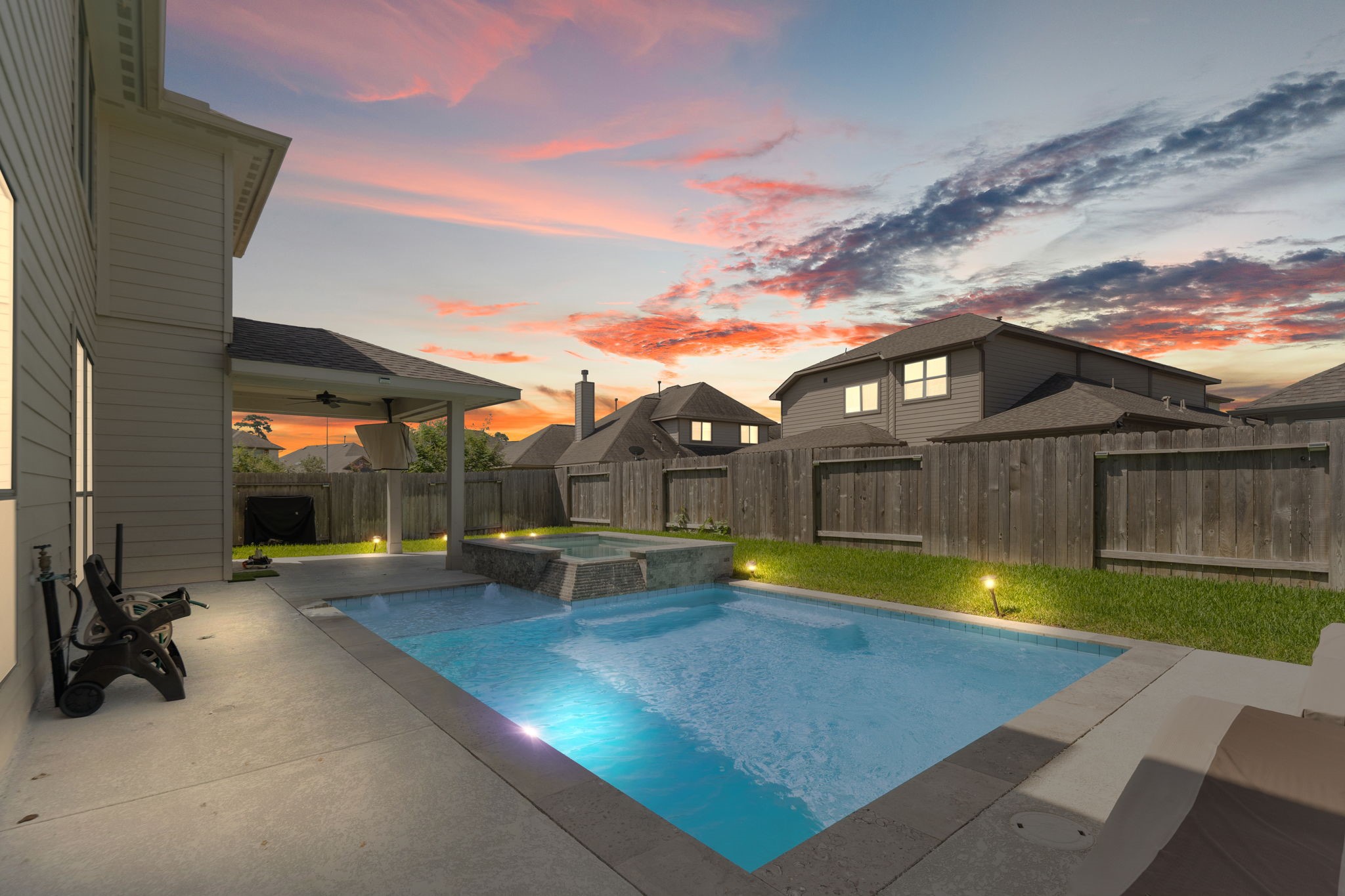 This backyard features a modern in-ground pool with integrated spa, surrounded by a spacious patio area. It offers a covered seating space, perfect for outdoor relaxation, against a backdrop of a scenic sunset sky.