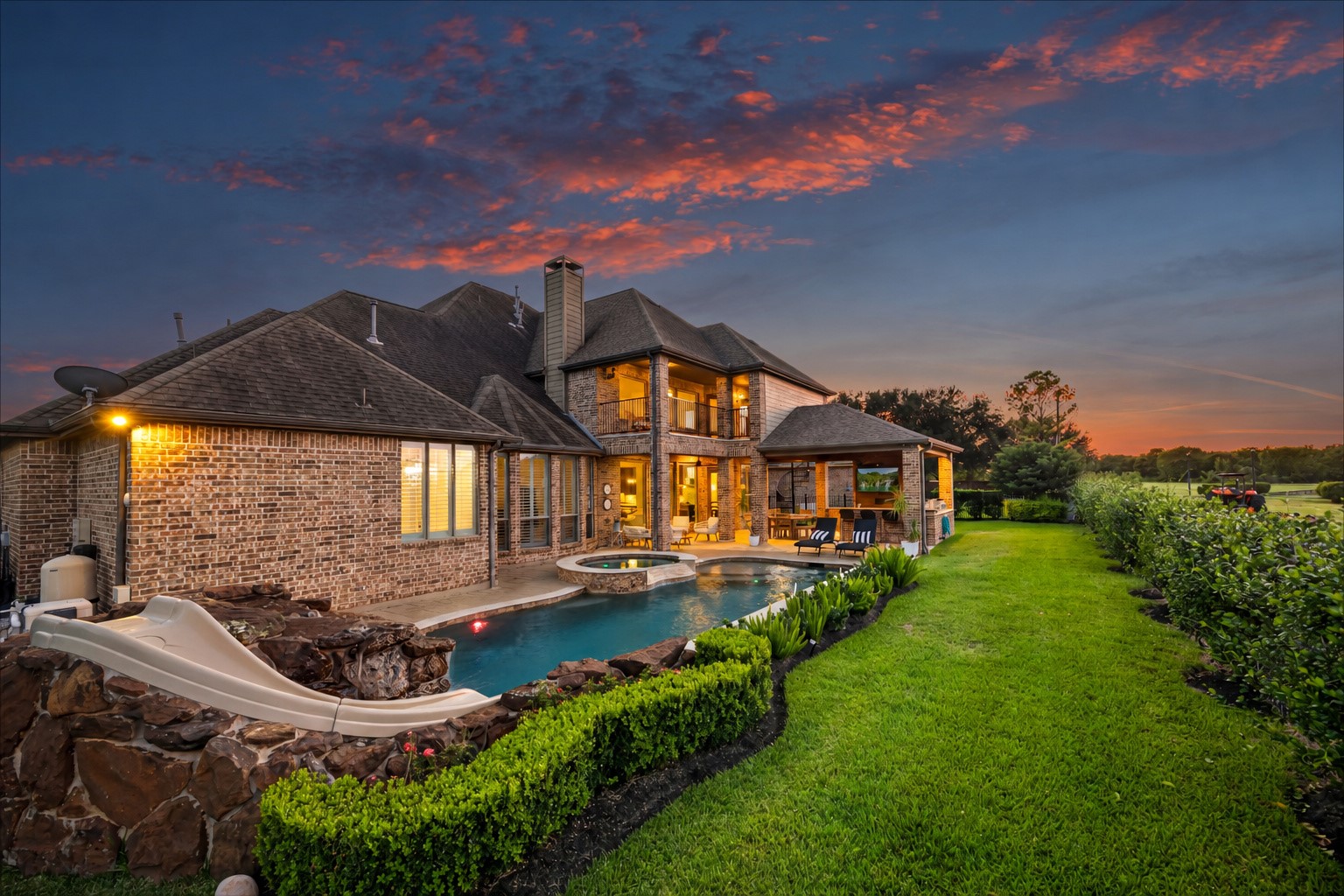 Resort-style backyard at dusk featuring a sparkling pool, outdoor kitchen, and expansive green space. Heated PebbleTec chlorine pool and outdoor kitchen with Alfresco grill and Alfresco flat grill.
