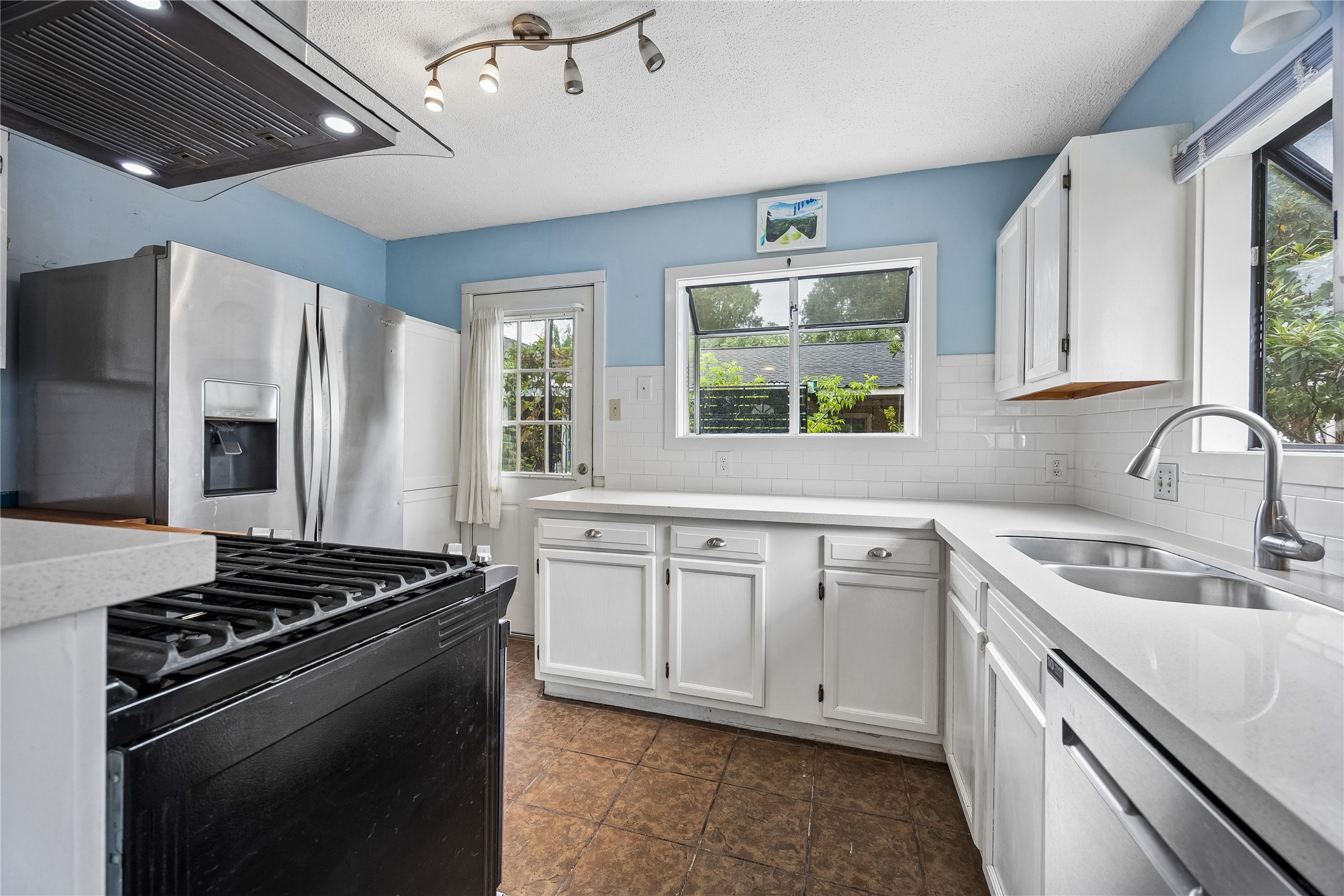 A bright, functional kitchen offers white cabinetry and countertops, stainless-steel refrigerator and dishwasher, a gas range, and windows overlooking the greenery outside