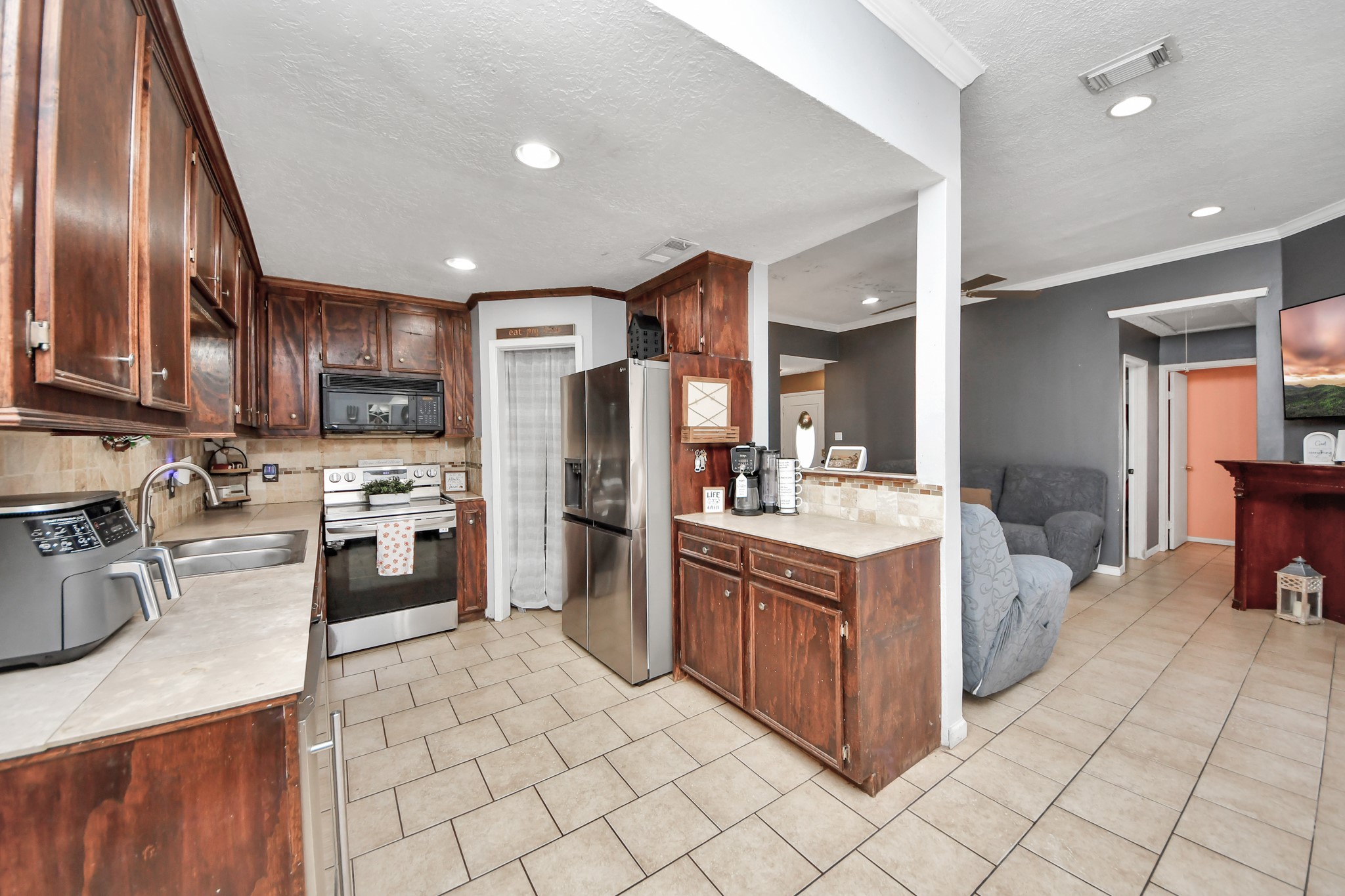 Spacious kitchen featuring rich wood cabinetry, tile flooring, ample countertop space, and a stylish tile backsplash. Complete with stainless steel appliances, a built-in wine cooler, and a convenient breakfast bar overlooking the living area