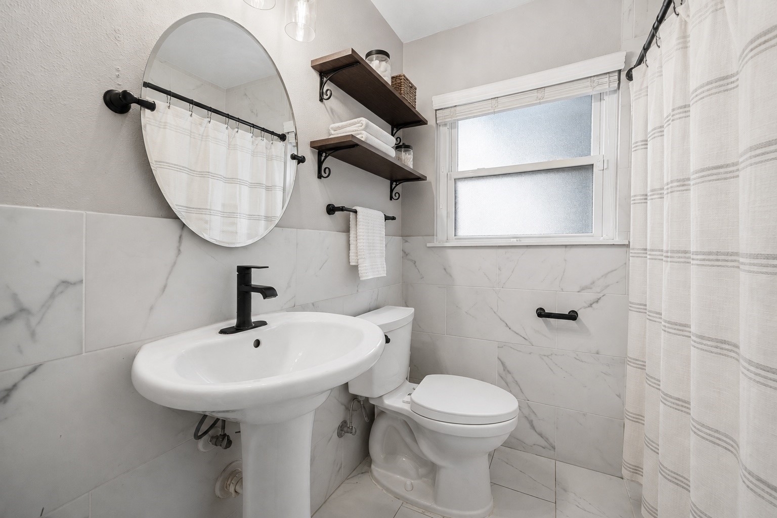 Fresh finishes give this bathroom a bright, updated feel, featuring marble look tile, matte black fixtures, open wood shelving, and a clean, timeless design. Decorative styling adds warmth while maintaining a crisp, spa inspired aesthetic. **Virtually staged**