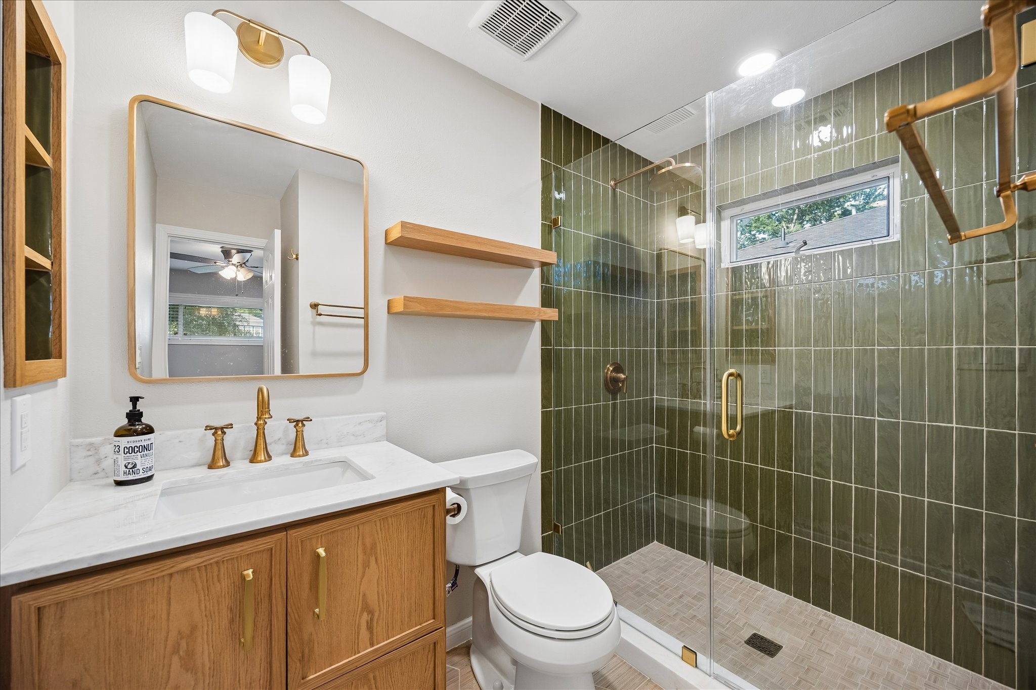 Rich green tile, warm wood cabinetry, and brushed brass fixtures create a striking designer aesthetic in the primary bath. A frameless glass shower, floating wood shelves, and marble look countertops blend timeless finishes with modern style. Rich green tile, warm wood cabinetry, and brushed brass fixtures create a striking designer aesthetic in the primary bath. A frameless glass shower, floating wood shelves, and marble look countertops blend timeless finishes with modern style.
