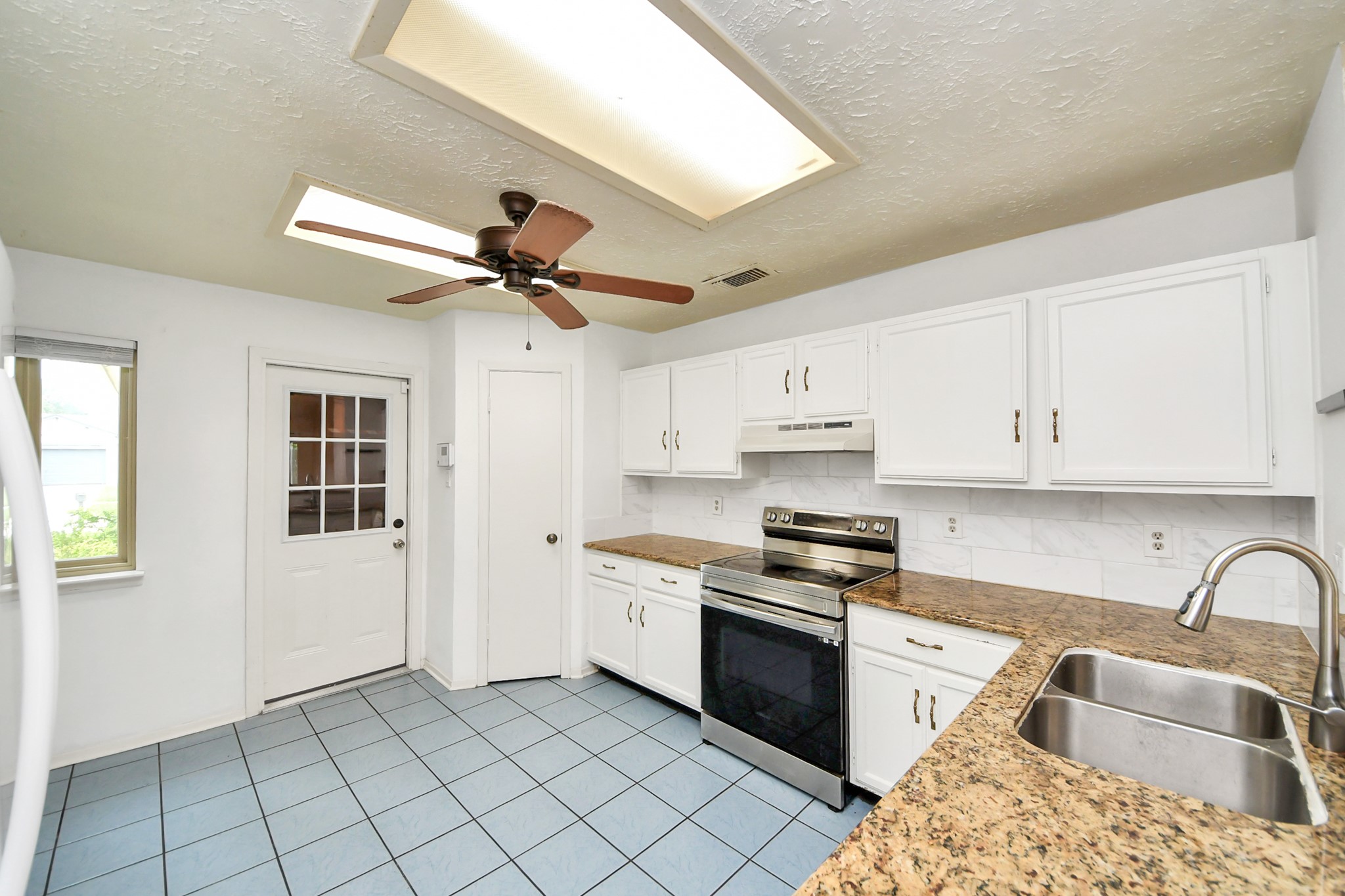 This bright kitchen features modern appliances, granite countertops, and ample white cabinetry. It includes a ceiling fan, tiled flooring, and a convenient door leading outside.