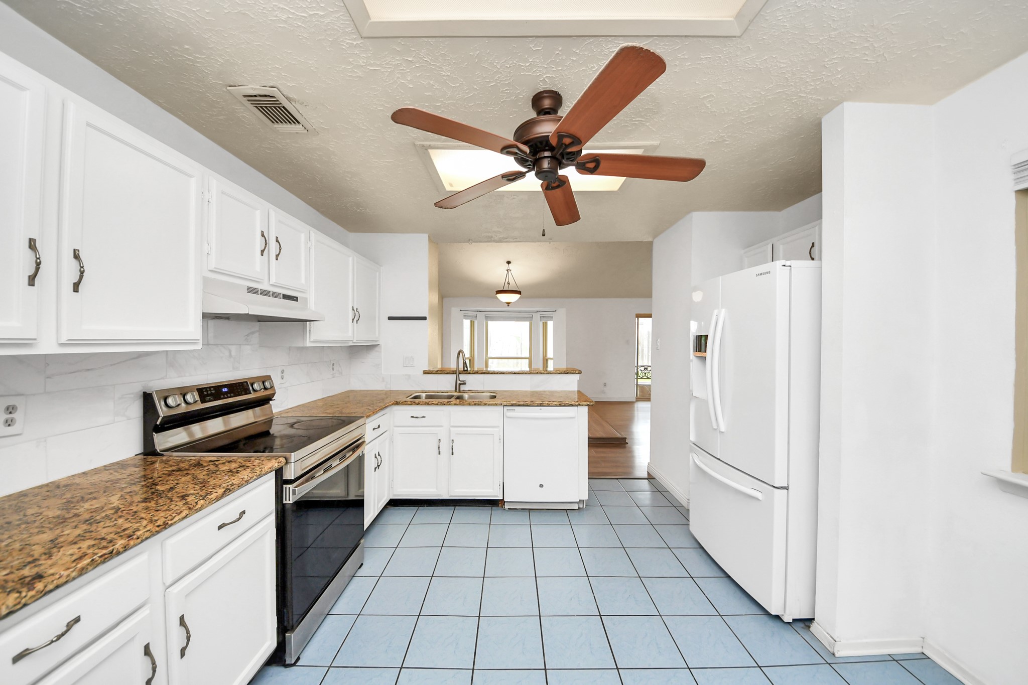 This kitchen features white cabinetry, granite countertops, and a modern stove. It has blue tile flooring and a large ceiling fan. The space opens into a dining area with ample natural light.