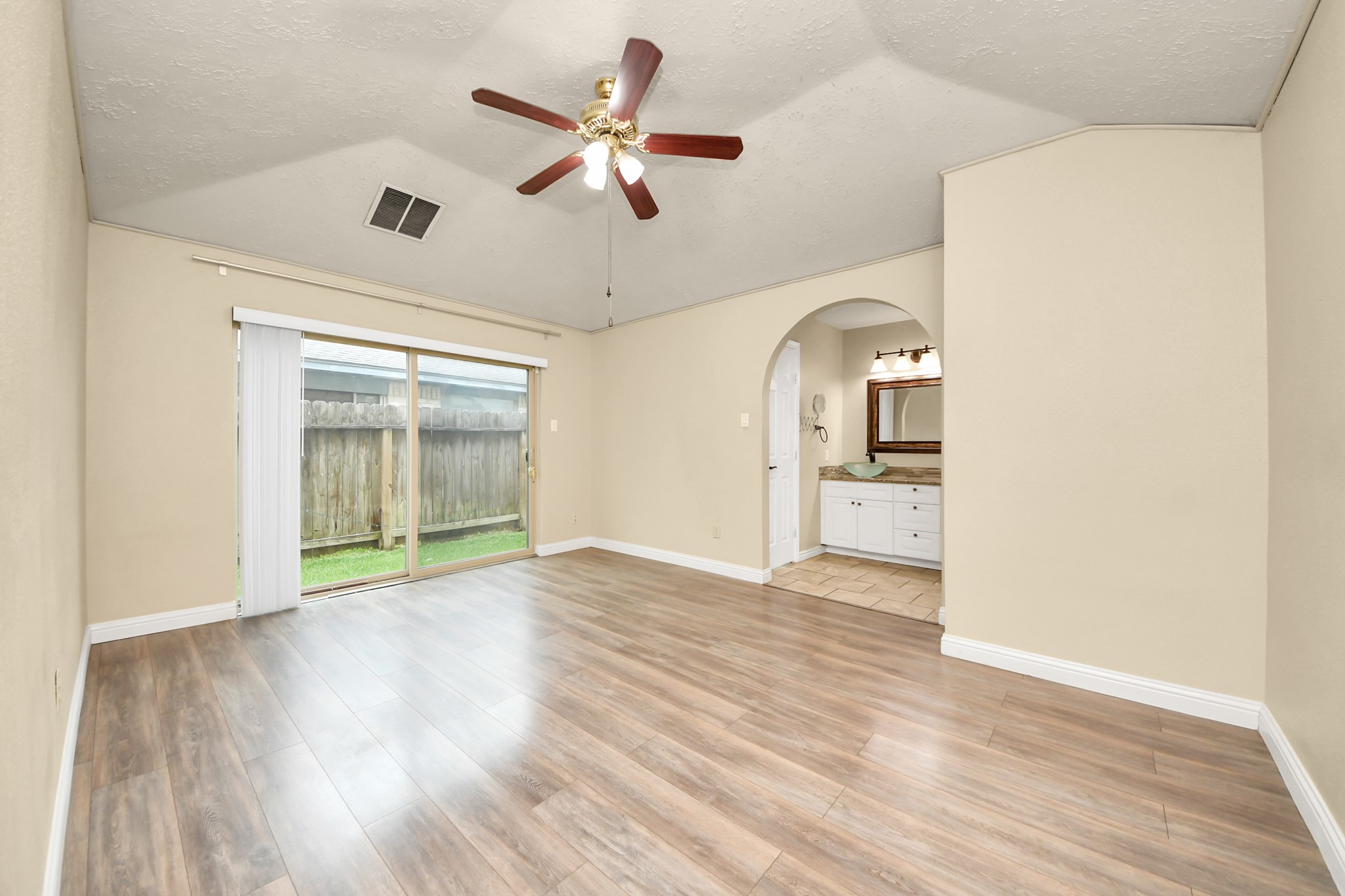 Bright room with vaulted ceiling, wood flooring, and a ceiling fan. Large sliding glass doors lead to a fenced backyard. An arched doorway opens to a bathroom with a modern sink and vanity.