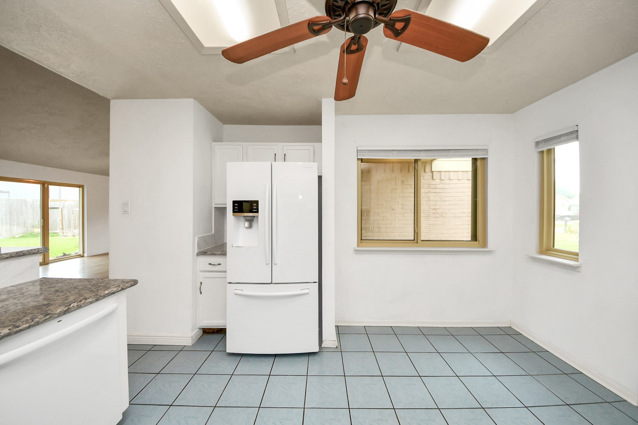 This kitchen area features light blue tile flooring, a ceiling fan, and ample natural light through two large windows. It includes a modern white refrigerator and a granite countertop, with a partial view of the backyard through sliding glass doors.