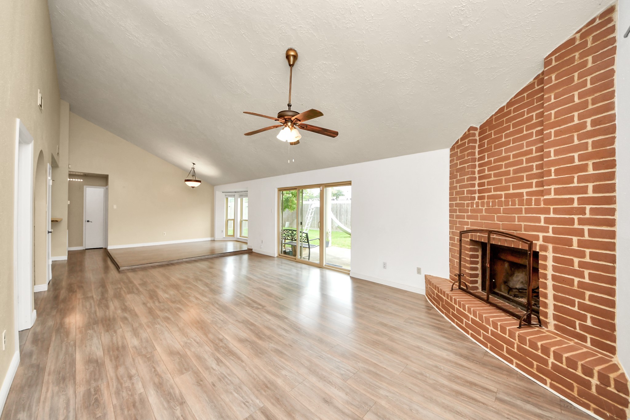 This spacious living area features a high vaulted ceiling, stylish wood flooring, and a striking brick fireplace. Natural light floods in through sliding glass doors, leading to a patio and backyard. Ideal for cozy gatherings and entertaining.