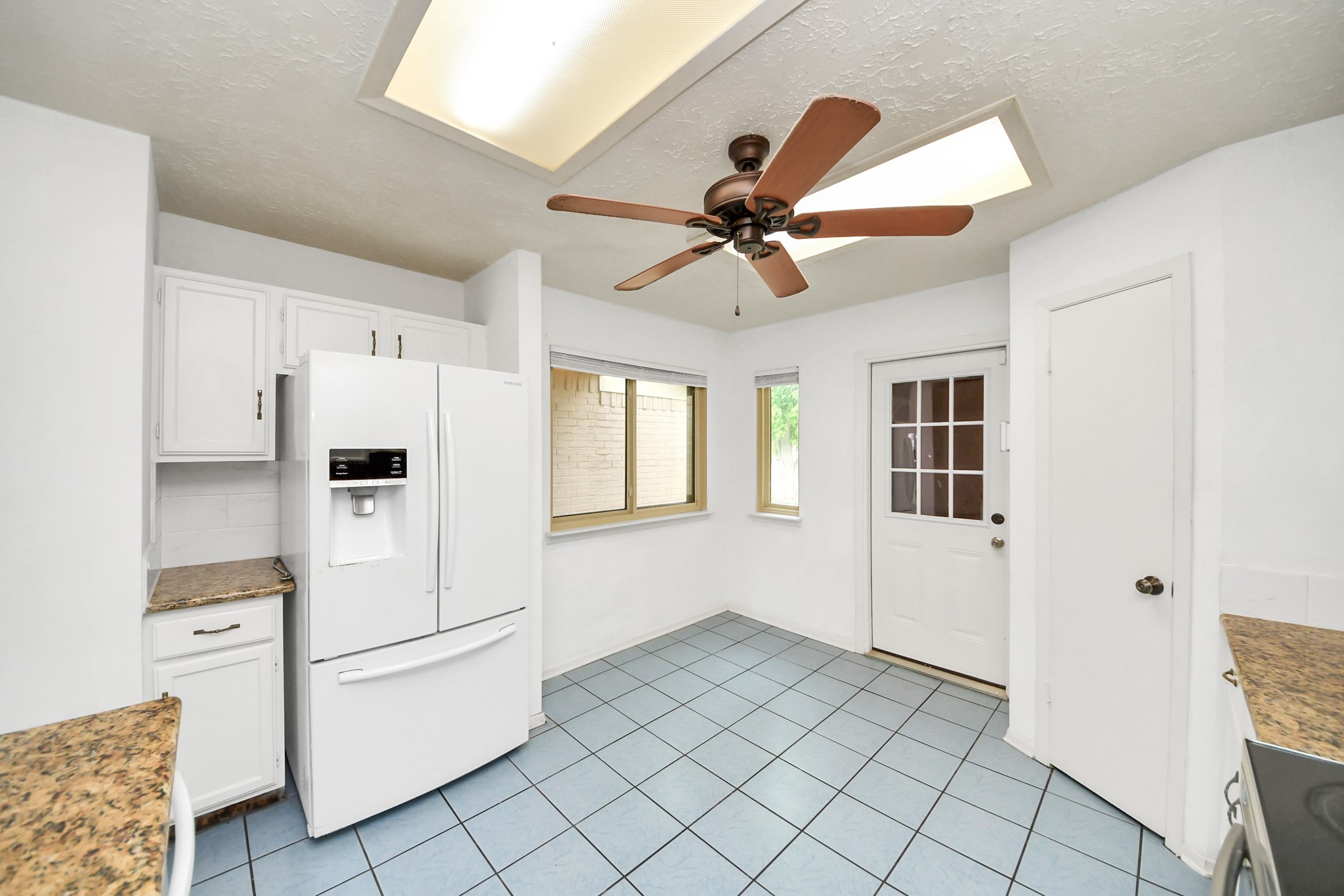 Bright kitchen with white cabinets, granite countertops, and a large refrigerator. Features blue tile flooring, a ceiling fan, and ample natural light from a window and a door with glass panels.
