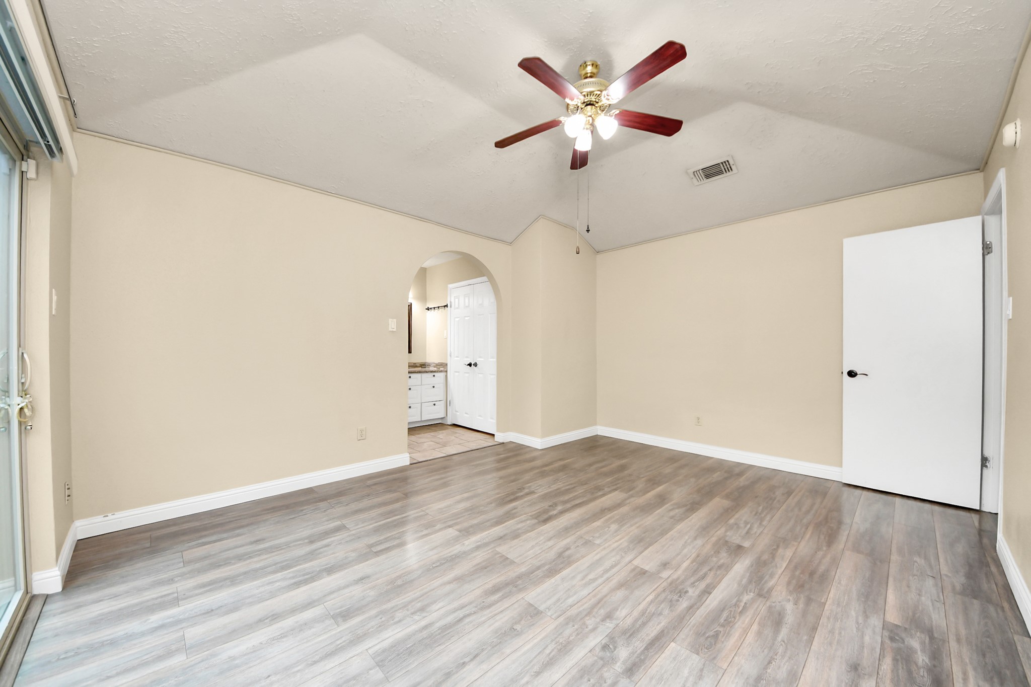 This room features light hardwood floors, neutral walls, and a ceiling fan. It has an arched doorway leading to a bathroom and large sliding glass doors that likely open to an outdoor space, providing ample natural light.