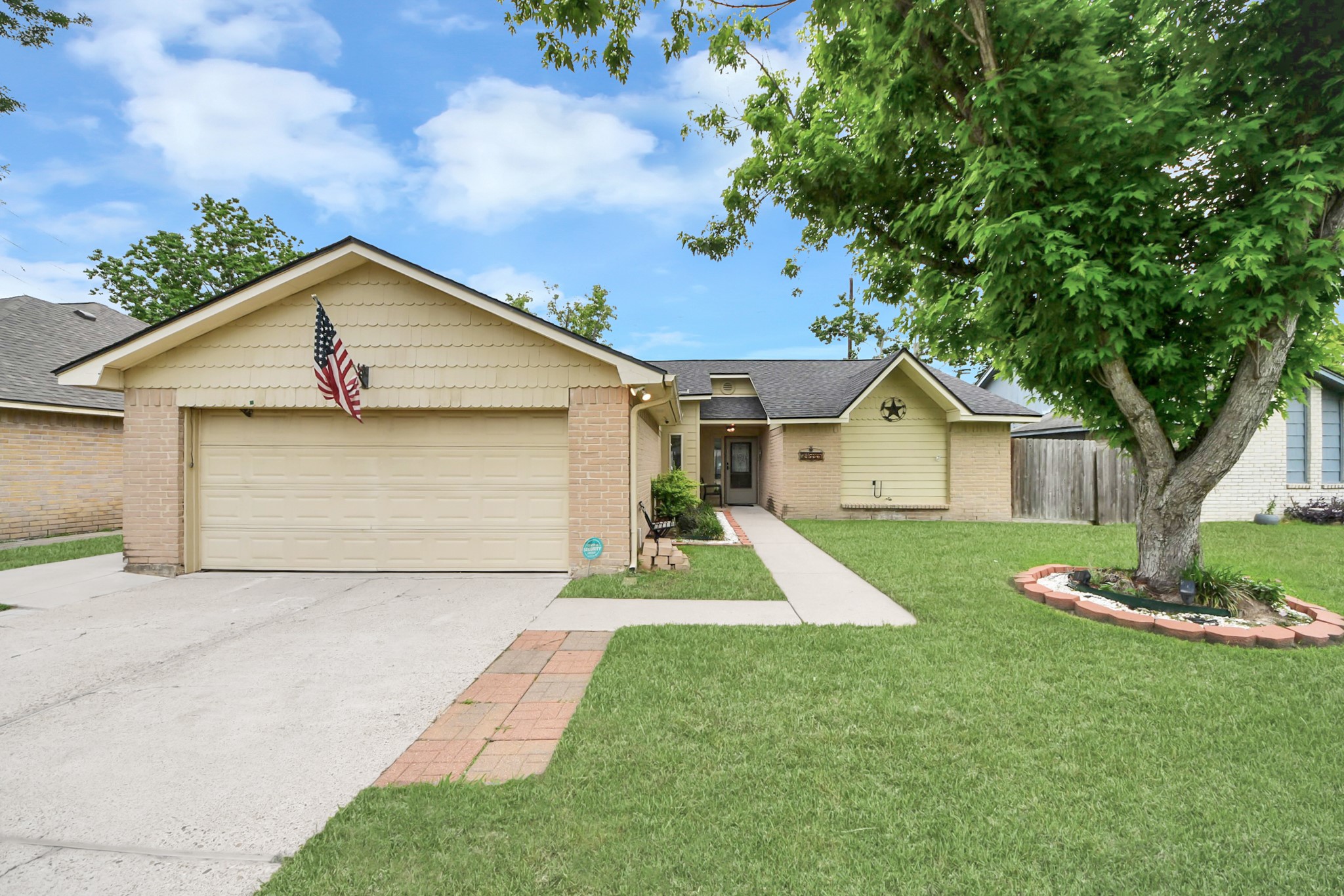 This inviting home features a charming exterior with a two-car garage, a neatly maintained front yard, and a large tree providing shade. A welcoming pathway leads to the entrance, making it an ideal choice for buyers seeking a cozy, suburban feel.