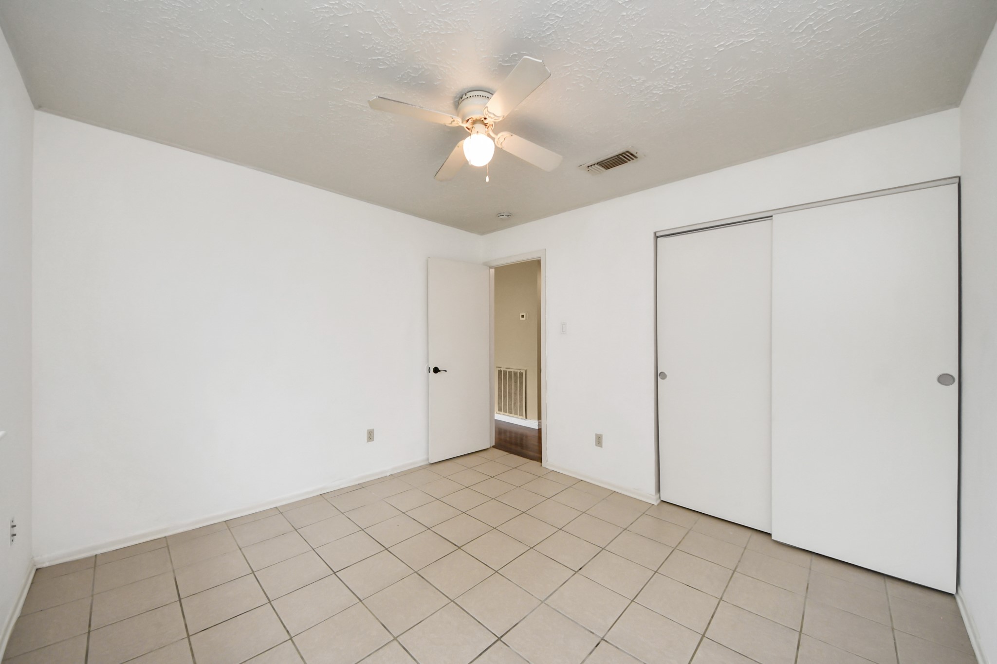 This room features a neutral color palette with white walls and tiled flooring. It includes a ceiling fan and a sliding door closet, providing functionality and storage.