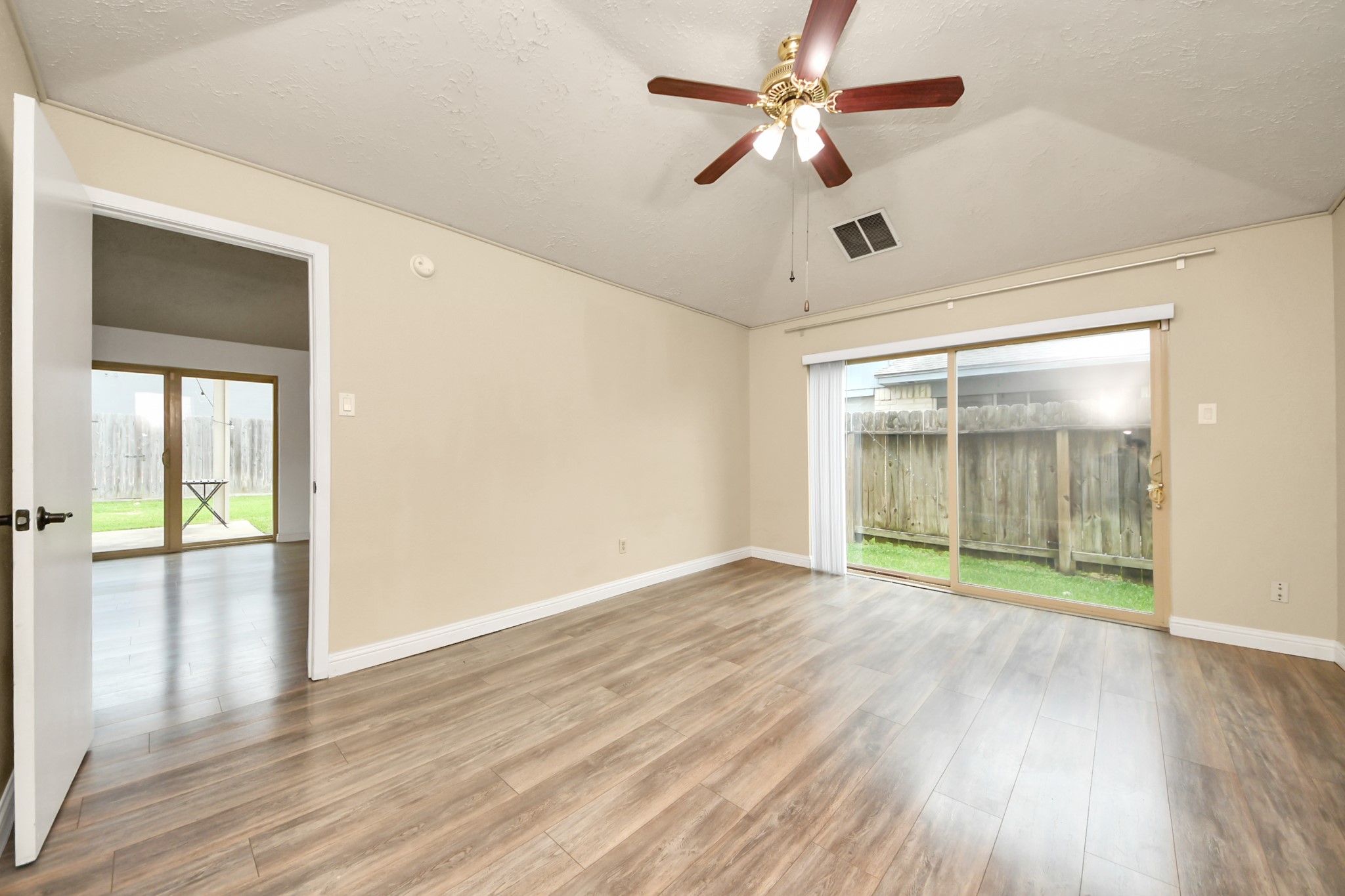 The primary bedroom Bright, spacious room with wood flooring, a ceiling fan, and large sliding glass doors leading to a fenced backyard. Natural light fills the space, creating an inviting atmosphere.
