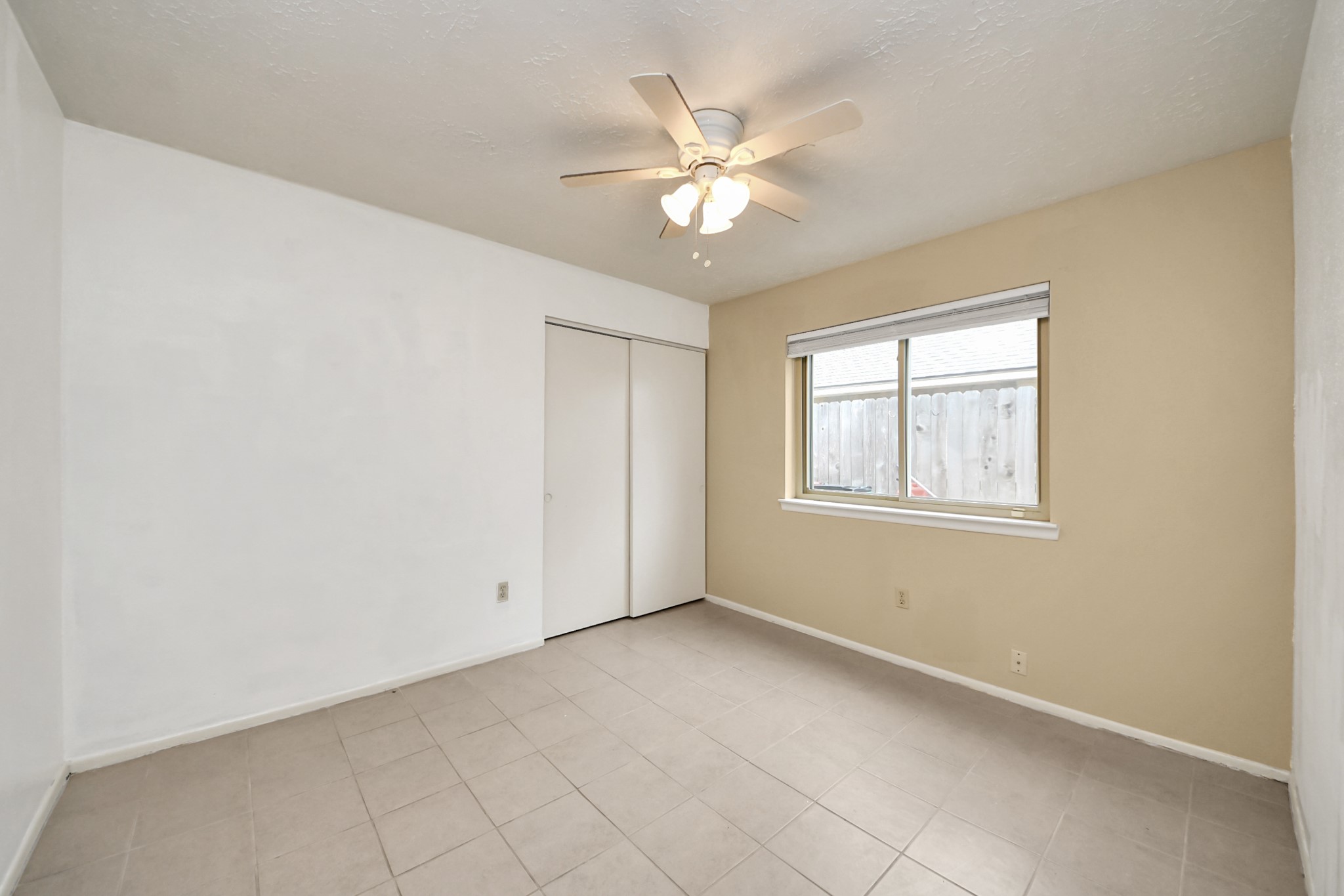 The bedroom features neutral tones with tiled flooring, a ceiling fan, and a large window providing natural light. It includes a closet with sliding doors, making it a versatile space for a bedroom or office.