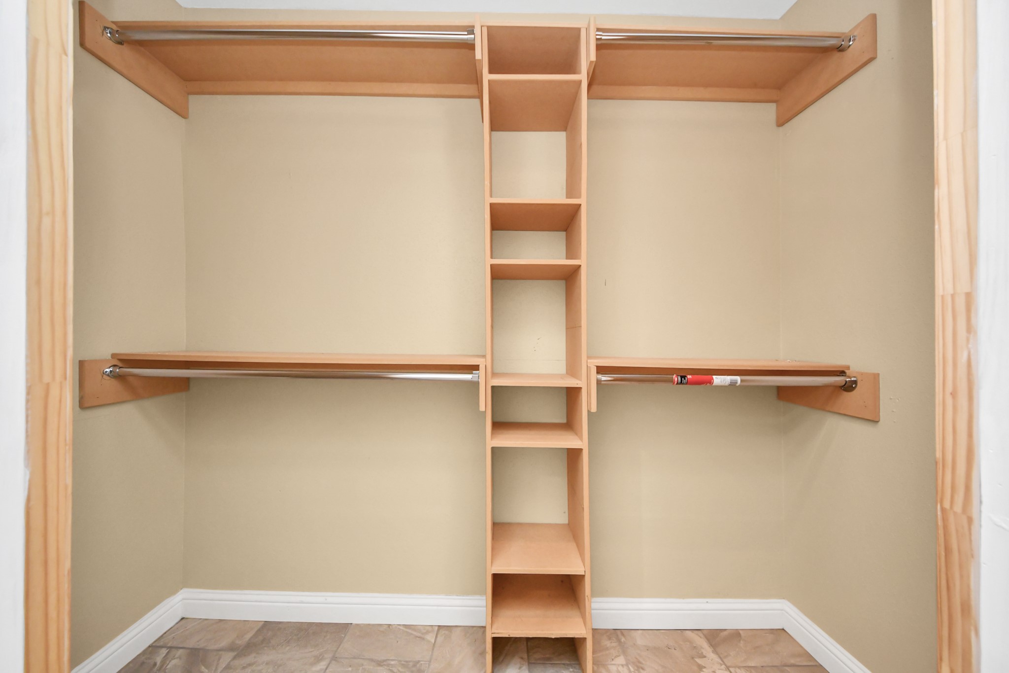 This is a spacious walk-in closet with a neutral color scheme, featuring multiple hanging rods and center shelving for organized storage. The tiled flooring adds a touch of elegance.
