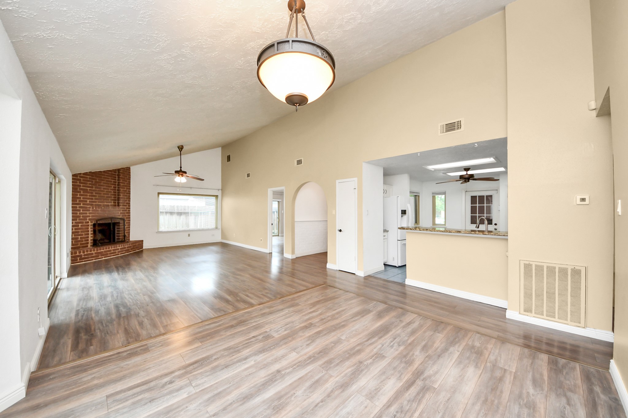 This spacious living area features high ceilings, wood flooring, and a cozy brick fireplace. It opens to a bright kitchen with a bar counter, perfect for entertaining. Large windows and sliding doors offer plenty of natural light.