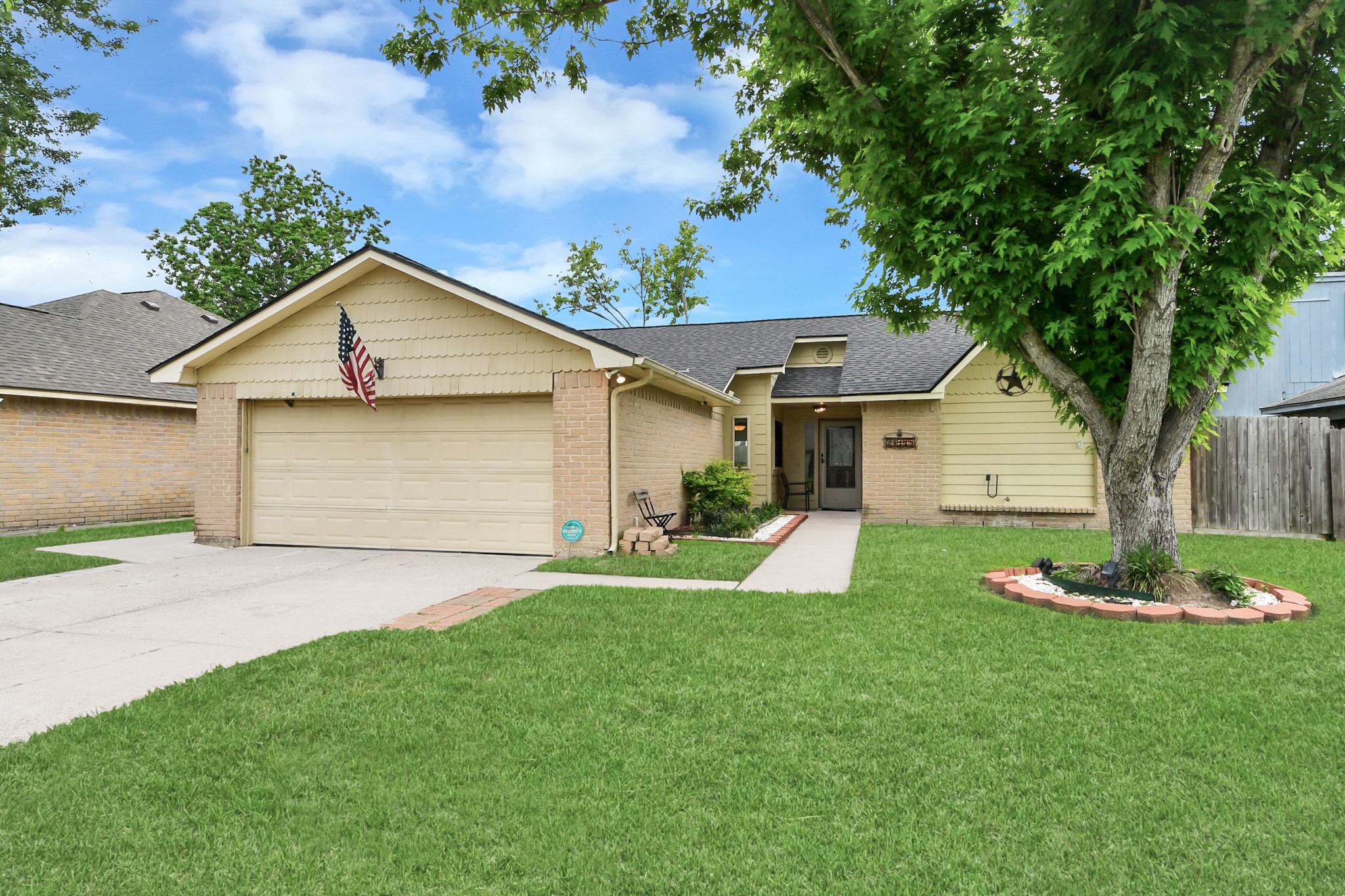 This charming single-story home features a well-maintained front yard with lush green grass, a mature tree, and a cozy porch. The exterior showcases a neutral color palette and a two-car garage, perfect for a welcoming suburban lifestyle.