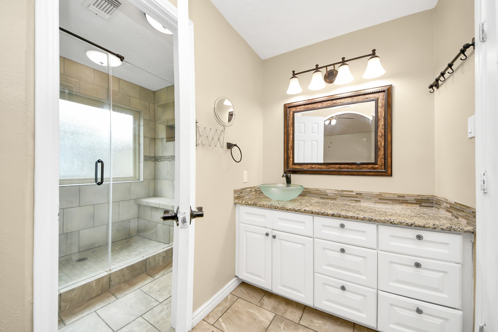 Primary bathroom features a spacious walk-in shower with glass doors, a sleek granite countertop with a modern vessel sink, and stylish lighting above a framed mirror. The neutral tile and cabinetry create a clean, elegant look.