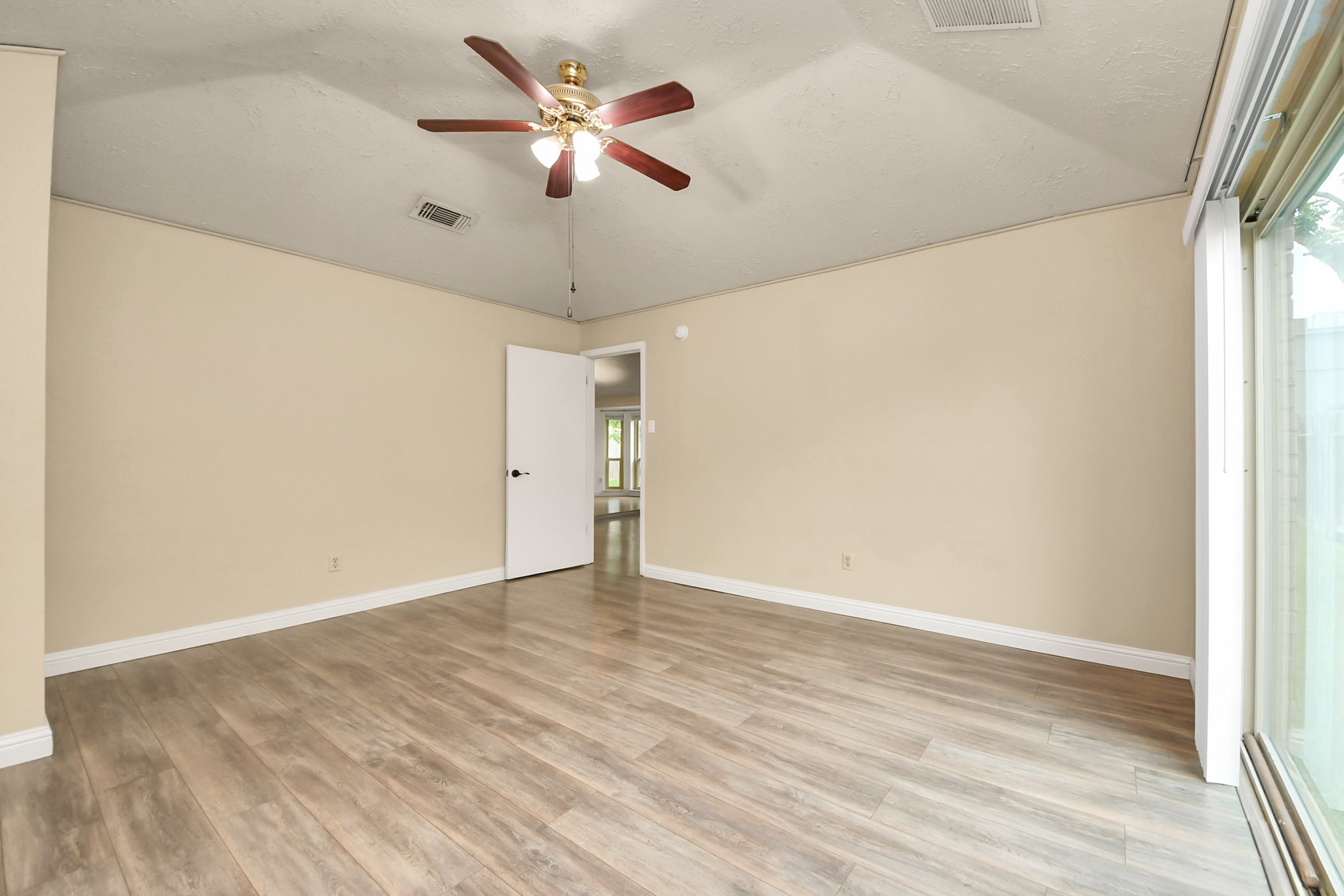 This spacious room features laminate wood flooring, neutral walls, a ceiling fan, and ample natural light from a large sliding glass door.