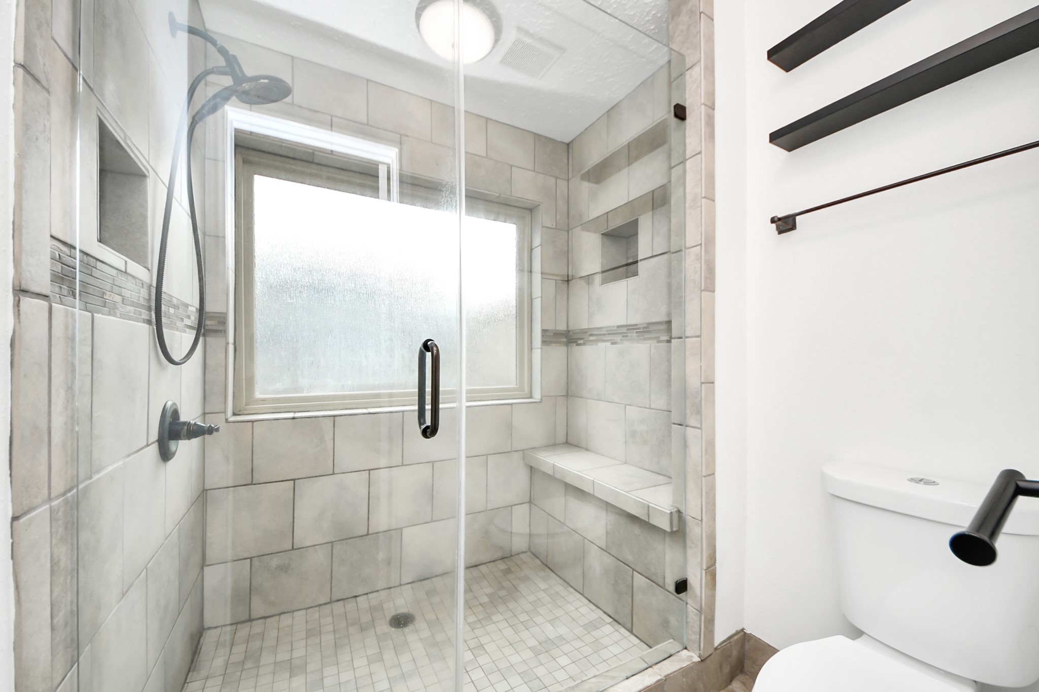 The primary bathroom features a modern walk-in shower with a glass door and a built-in bench. It has tiled walls and floor, a large window for natural light, and sleek fixtures for a clean, contemporary look.