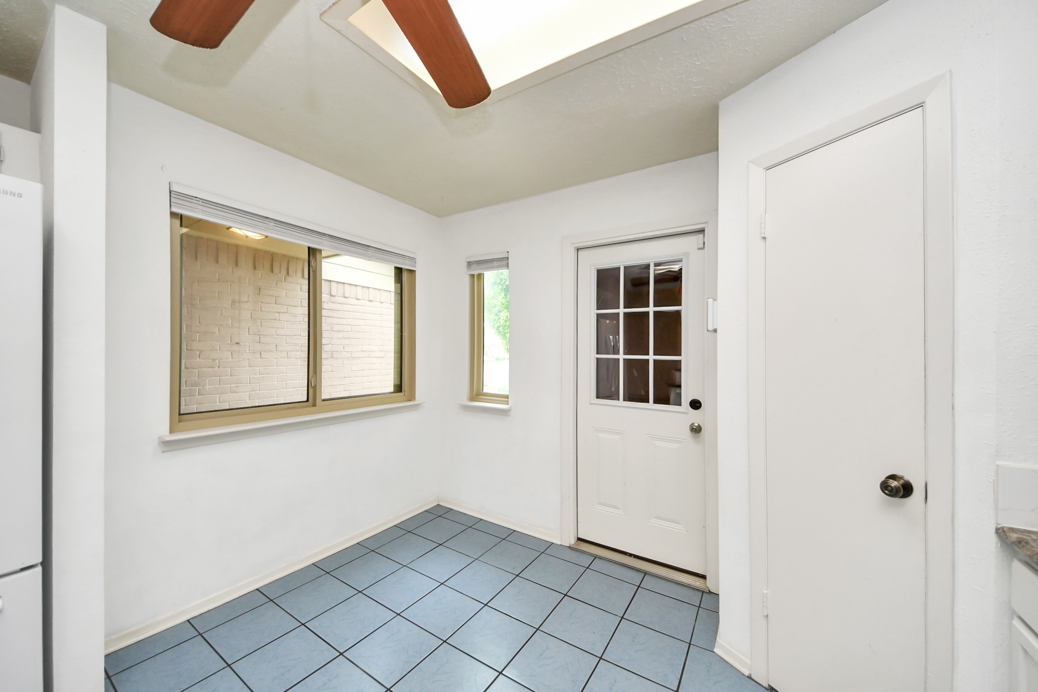 This bright room features a skylight, blue-tiled floor, and natural light from two windows. It includes a door with a glass panel leading outside and a white door to the side, offering a clean and versatile space.