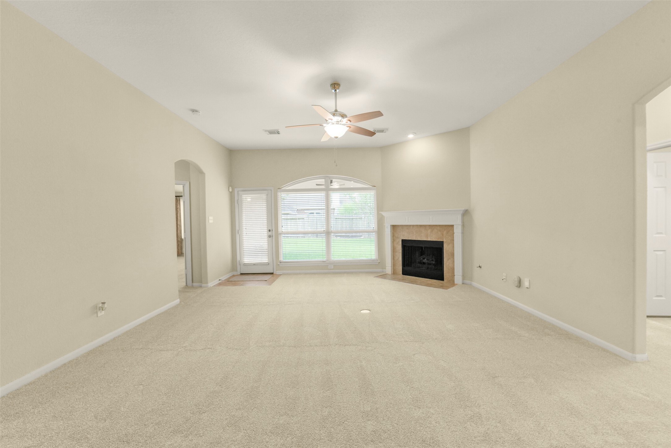 Spacious living room with plush carpeting, a cozy fireplace, large windows for natural light, and a ceiling fan, offering comfort and style.