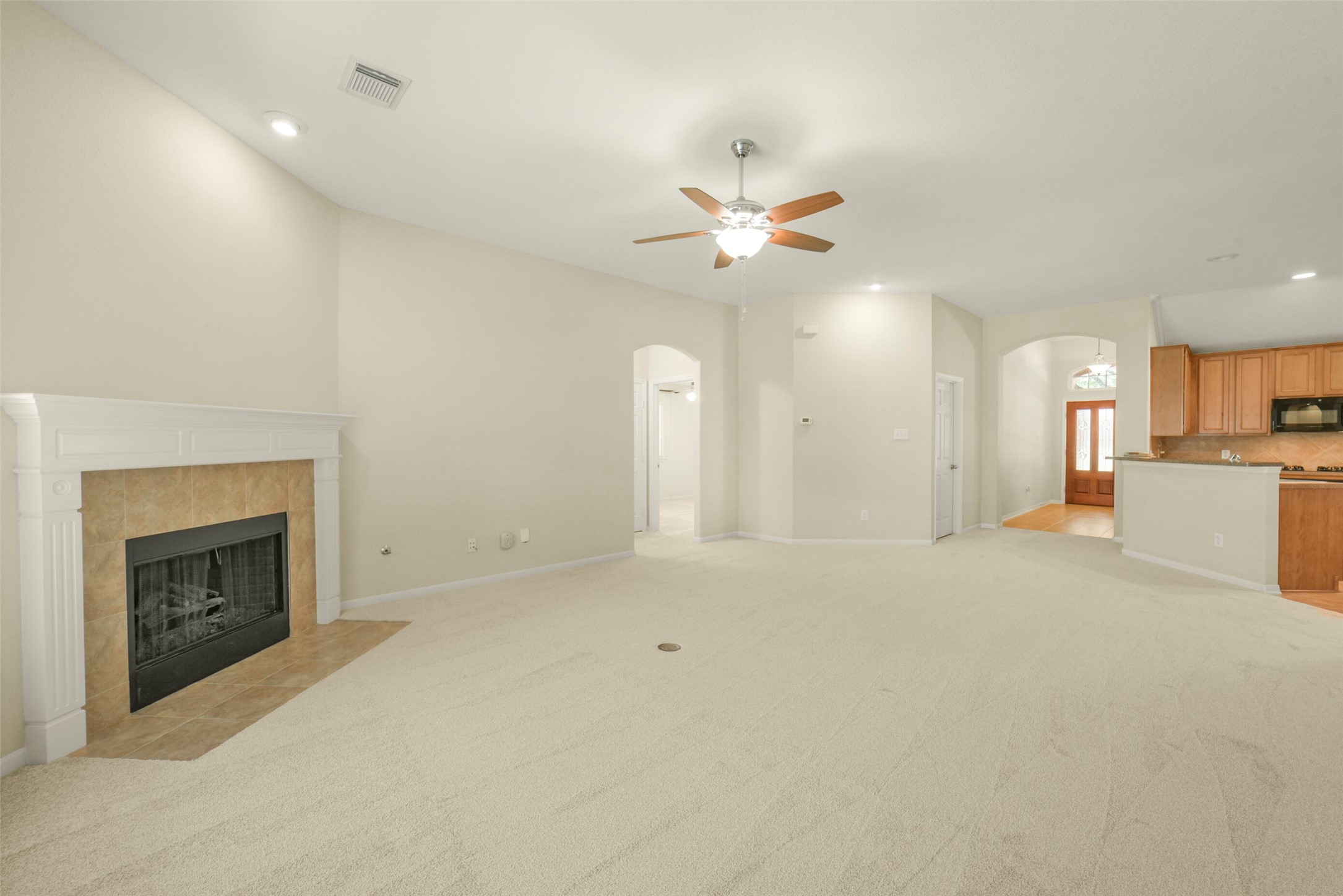 Spacious living room with plush carpeting, a cozy fireplace, and an open layout leading to a modern kitchen with wood cabinetry.