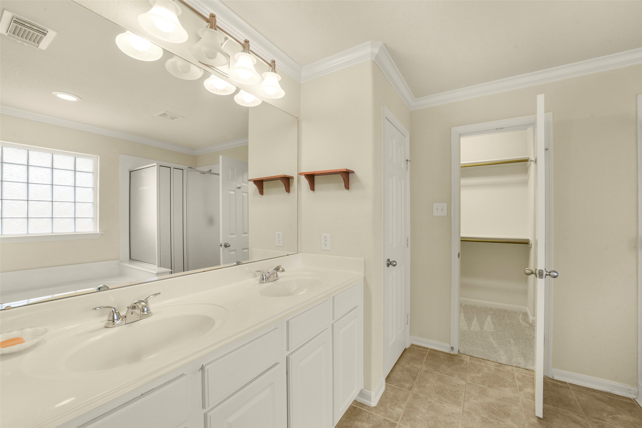 Spacious primary bathroom featuring a dual-sink vanity, glass-enclosed shower, and a large walk-in closet, all illuminated by natural light from a frosted window.