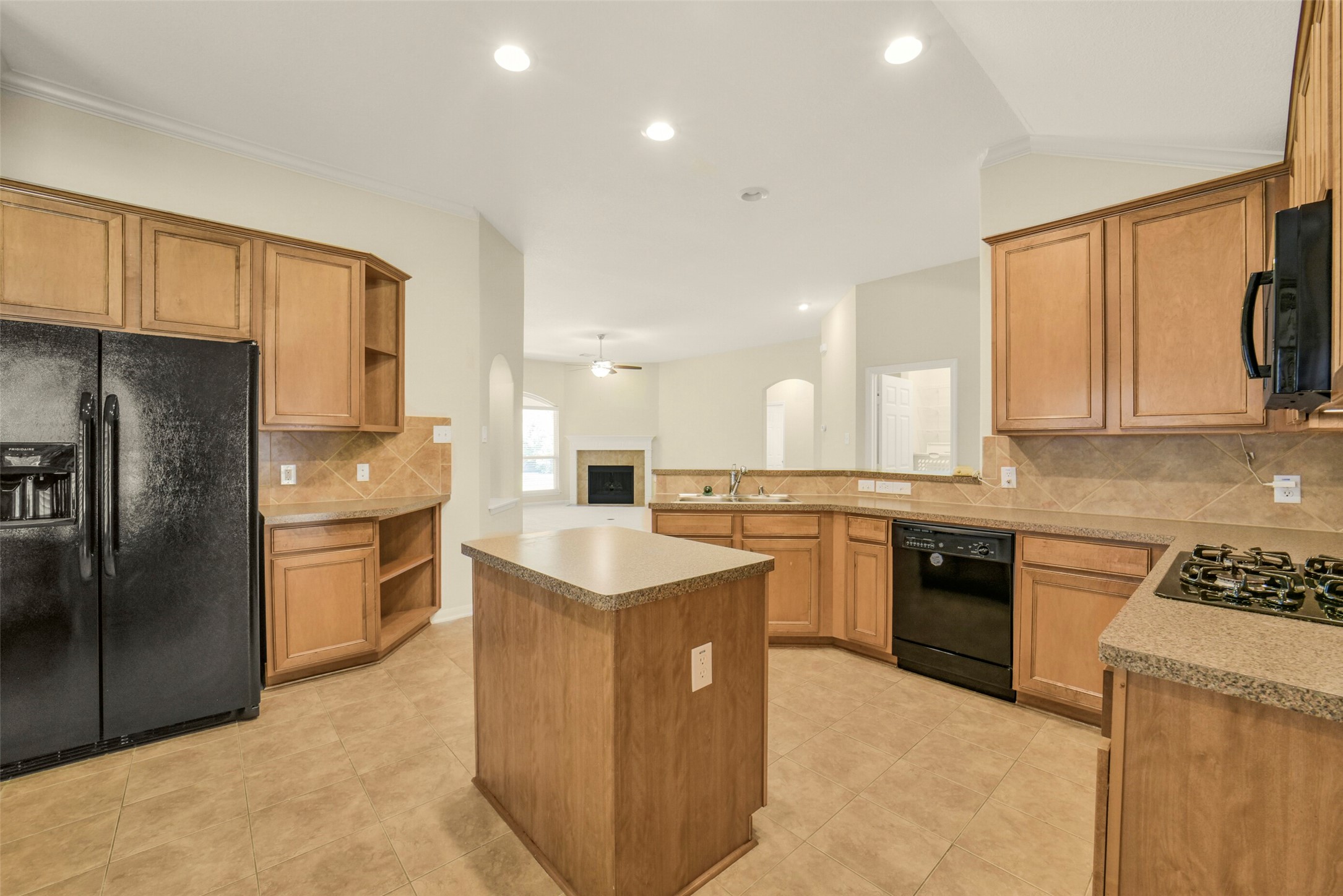 Spacious kitchen featuring warm wood cabinetry, sleek black appliances, and a central island, flowing seamlessly into a bright living area with a cozy fireplace.