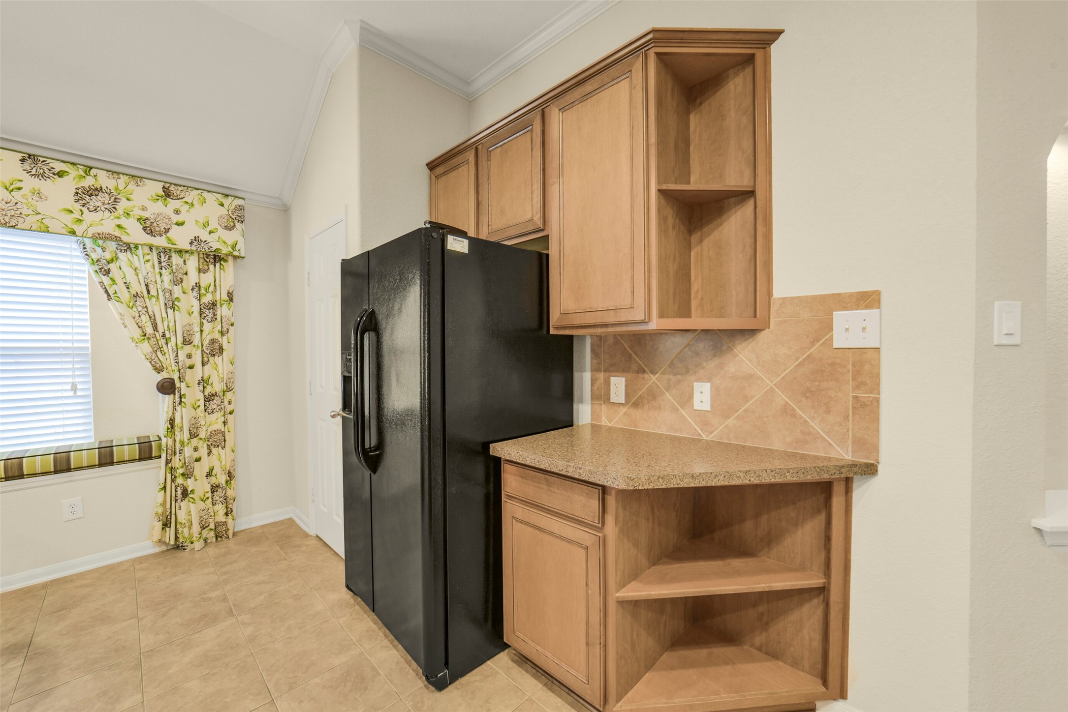 Charming kitchen corner with light wood cabinetry, sleek black refrigerator, and a cozy window seat adorned with floral drapes.