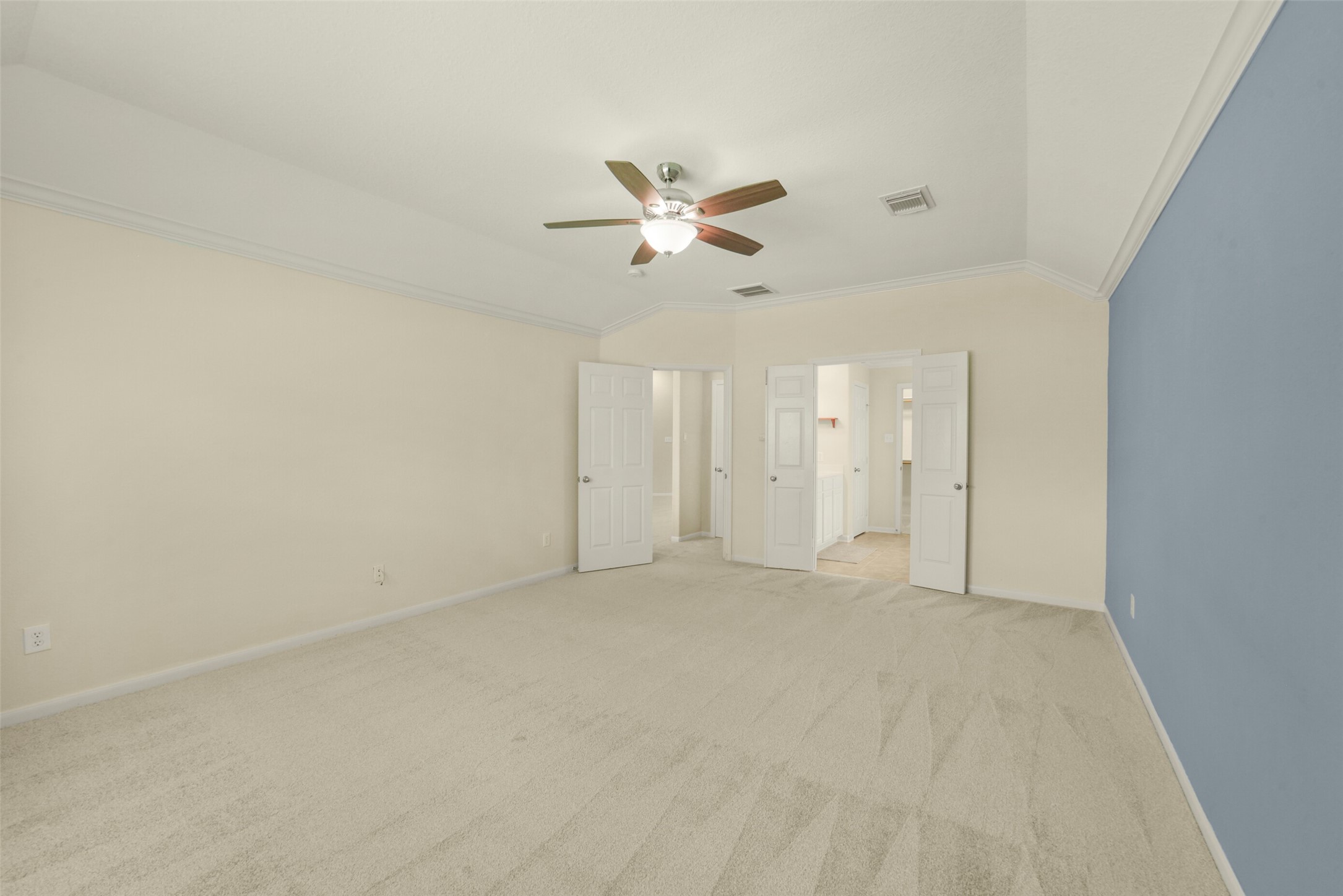 Spacious primary bedroom featuring plush carpeting, a soothing blue accent wall, and a ceiling fan, leading to an en-suite bathroom through double doors.