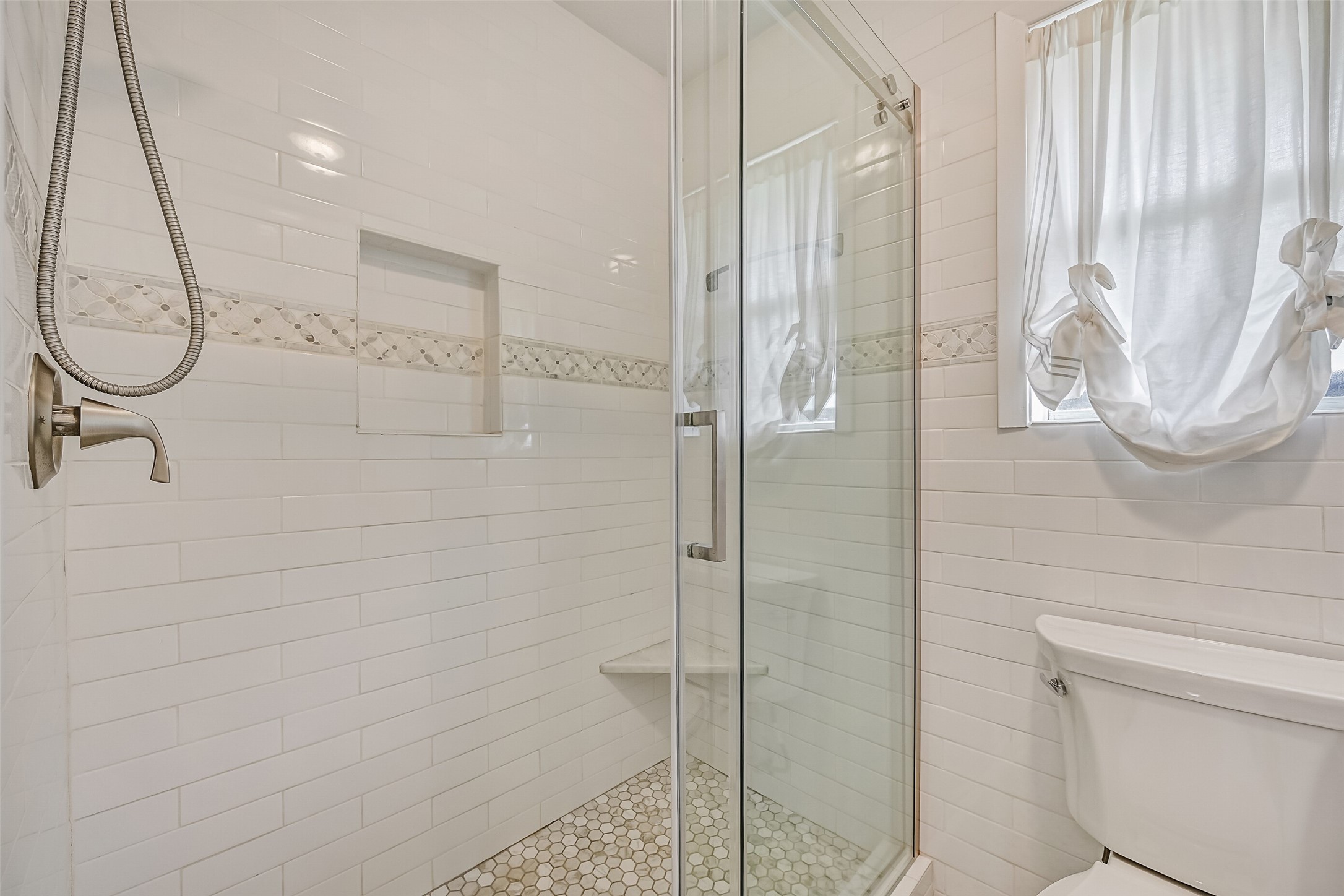 Master Bathroom  First Residence:
Walk-in glass-enclosed shower featuring classic white subway tile, a decorative mosaic accent band, built-in niche and bench, hex tile flooring, and a window providing natural light for a bright, spa-inspired feel.