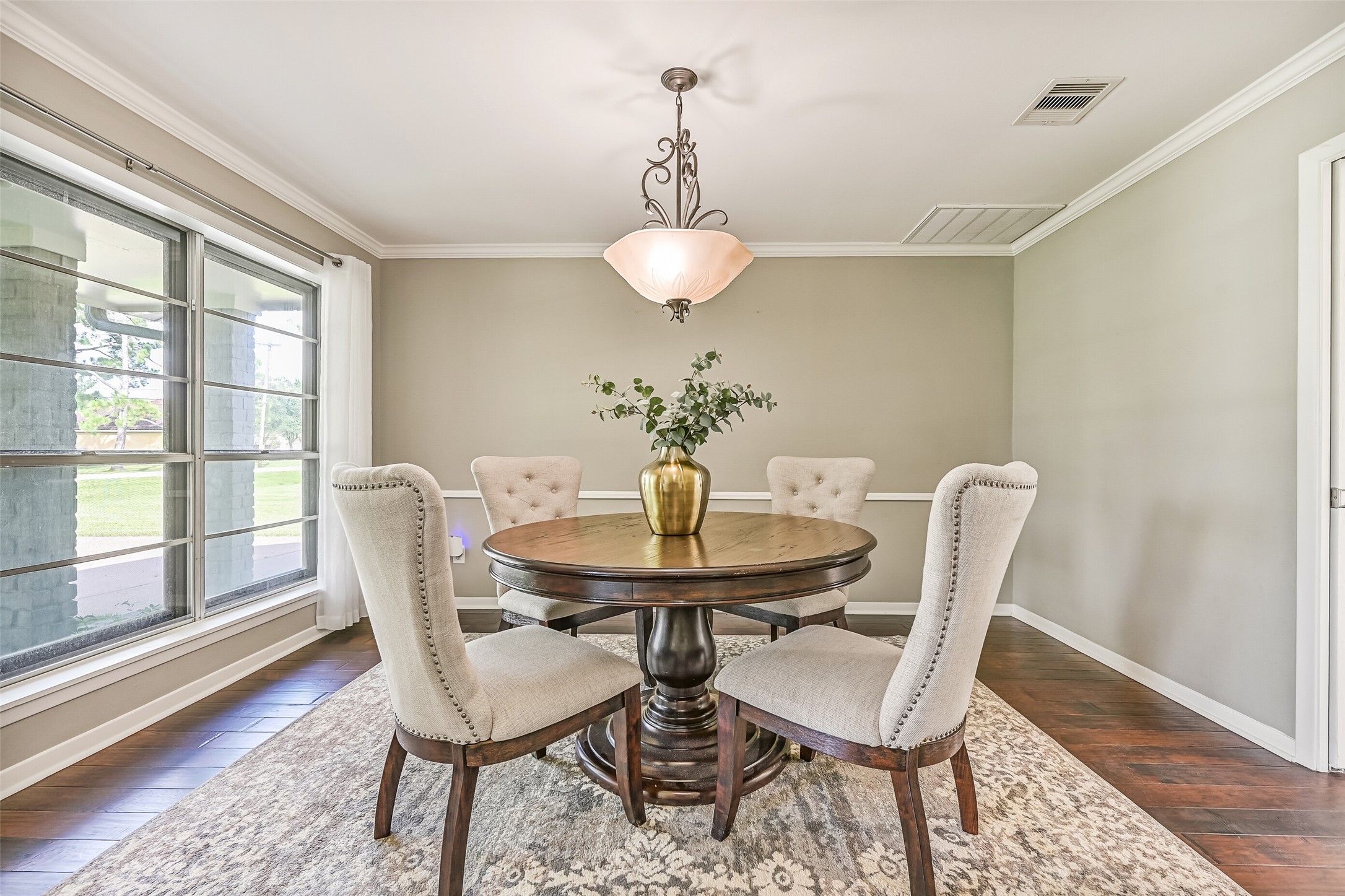 Dining Room  First Residence:
Formal dining room featuring wood flooring, crown molding, and a dimmable pendant light fixture, with large windows and breezy draperies showcasing views of the front porch and yard.