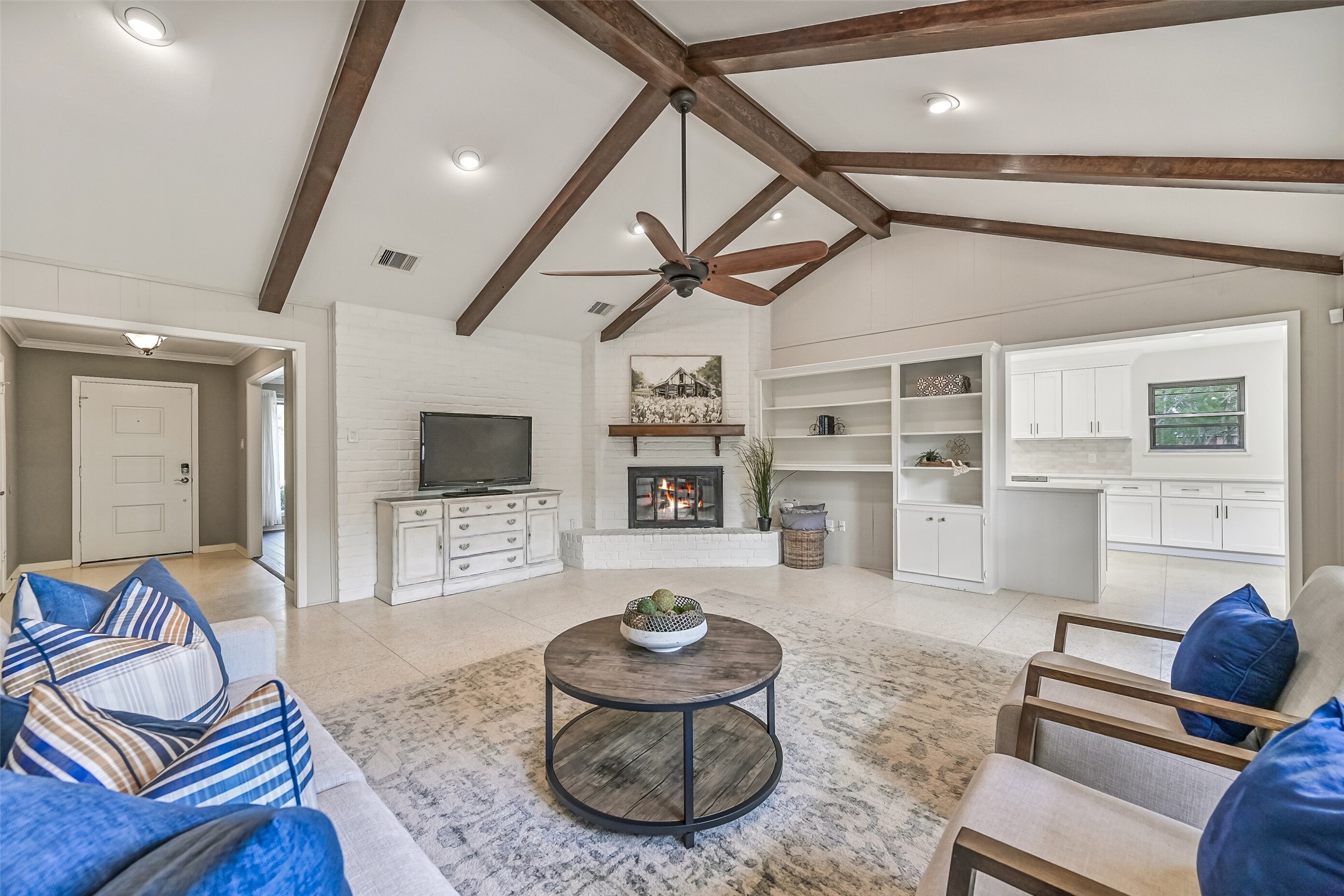 Family Room  First Residence:
Step from the foyer into the spacious family room highlighted by a vaulted ceiling with exposed wood beams, recessed lighting, and a ceiling fan, anchored by a painted brick corner-set gas log fireplace and retro terrazzo tile flooring.