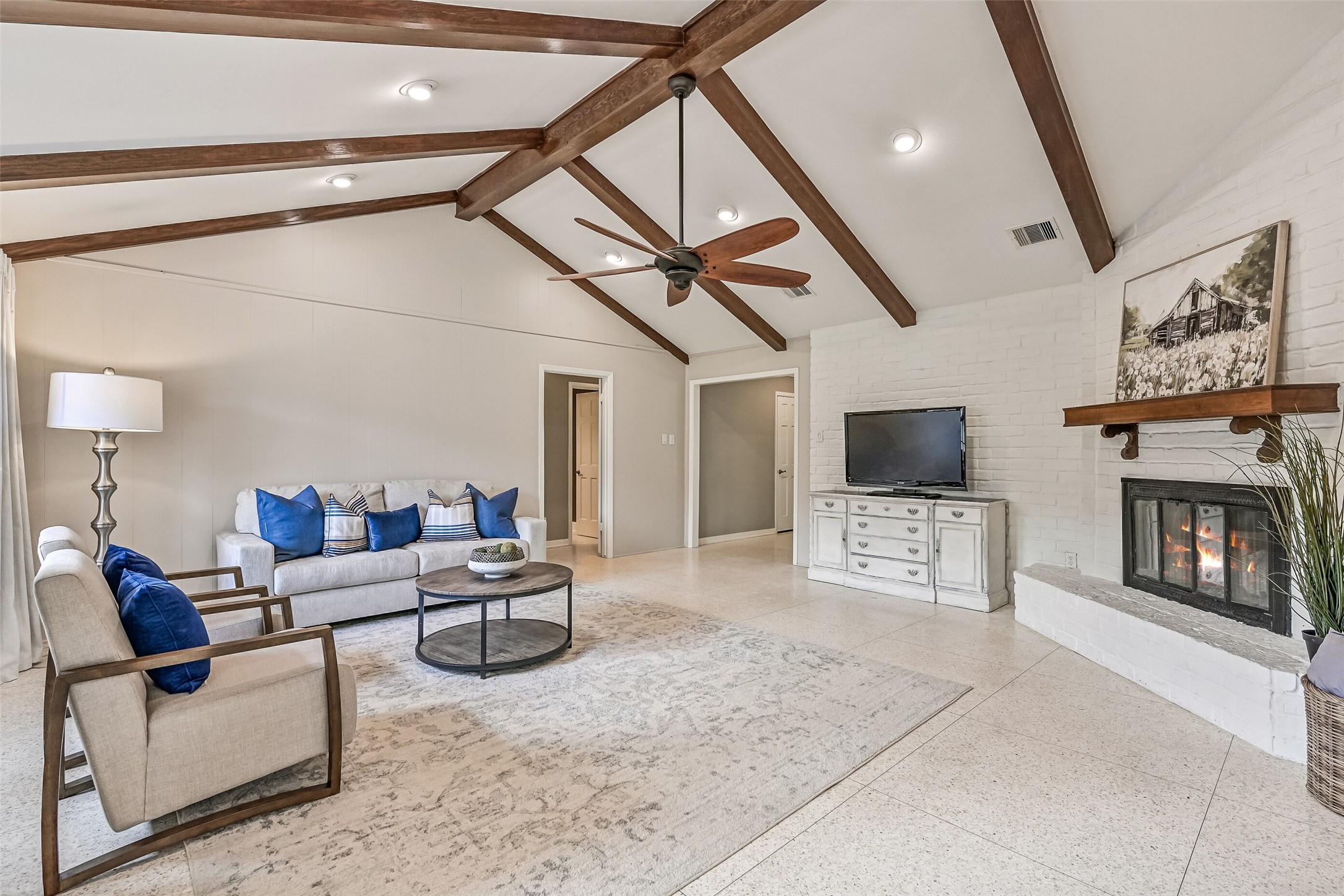Family Room  First Residence:
Step from the foyer into the spacious family room highlighted by a vaulted ceiling with exposed wood beams, recessed lighting, and a ceiling fan, anchored by a painted brick corner-set gas log fireplace and retro terrazzo tile flooring.
