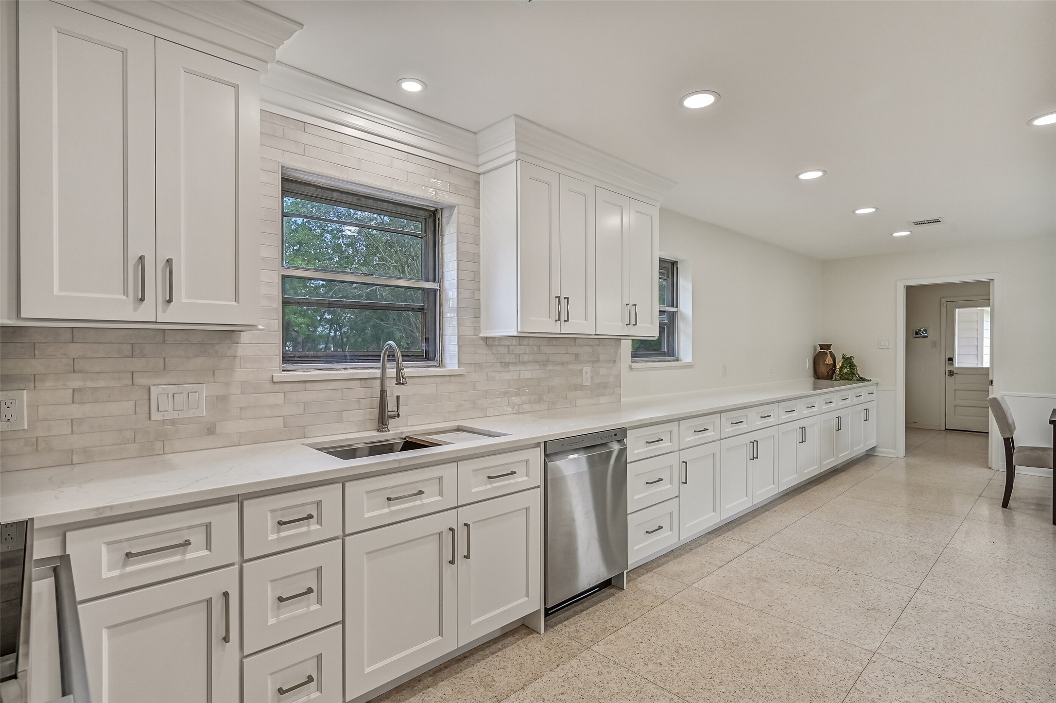 Kitchen  First Residence:
This view of the kitchen workspace highlights expansive quartz countertops, a dual basin sink positioned below a sunlit window, the new stainless dishwasher, complemented by abundant cabinetry and generous prep and serving surfaces extending into the breakfast room.