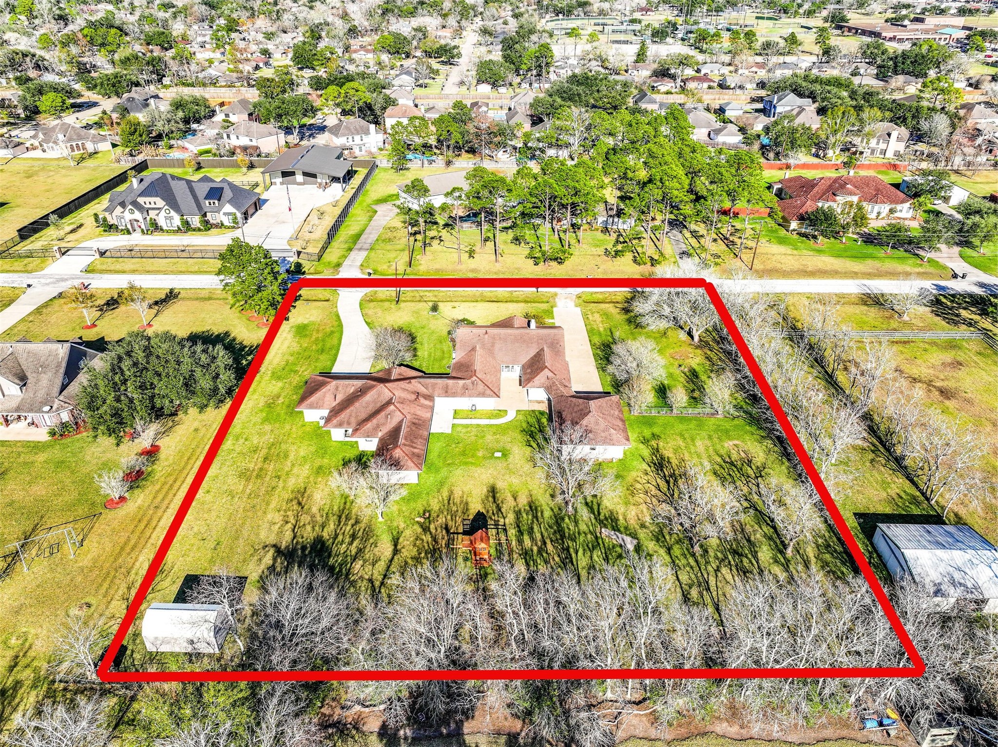 Expansive 1.3-acre Friendswood property boasts a unique dual-residence layout, featuring two complete homes with individual driveways, surrounded by mature trees & wide-open green space. Aerial views showcase the generous lot size with shed, playground, finished garage space, & desirable location on a quiet street with no HOA!