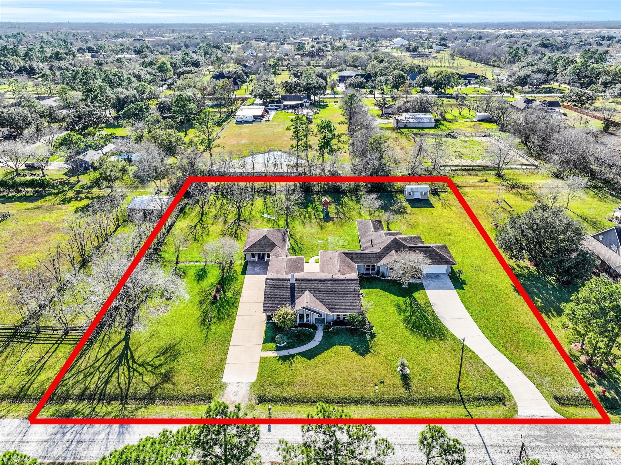 Expansive 1.3-acre Friendswood property boasts a unique dual-residence layout, featuring two complete homes with individual driveways, surrounded by mature trees & wide-open green space. Aerial views showcase the generous lot size with shed, playground, finished garage space, & desirable location on a quiet street with no HOA!