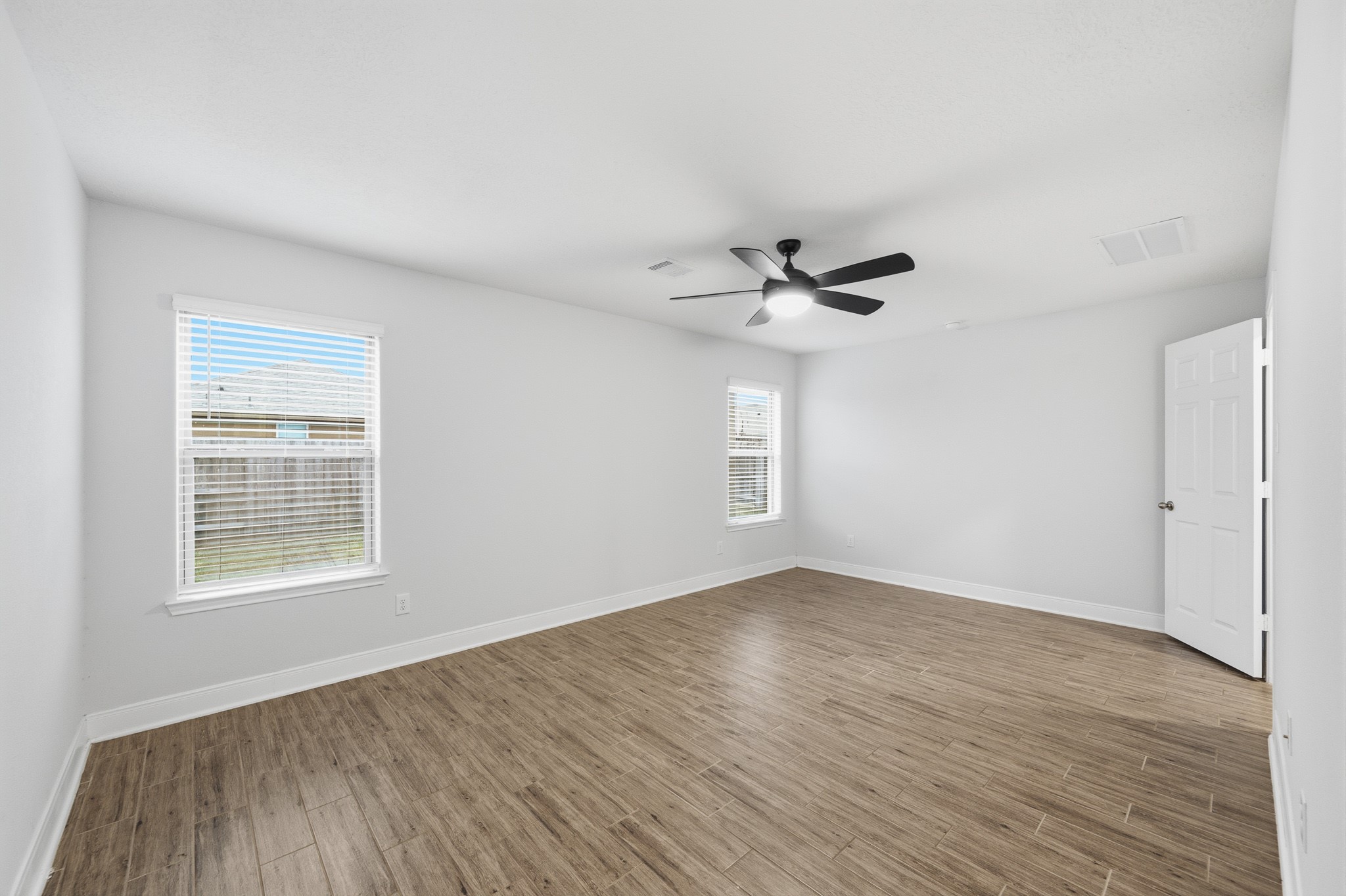 Bright and spacious room featuring sleek flooring, a modern ceiling fan, and ample natural light from two large windows.