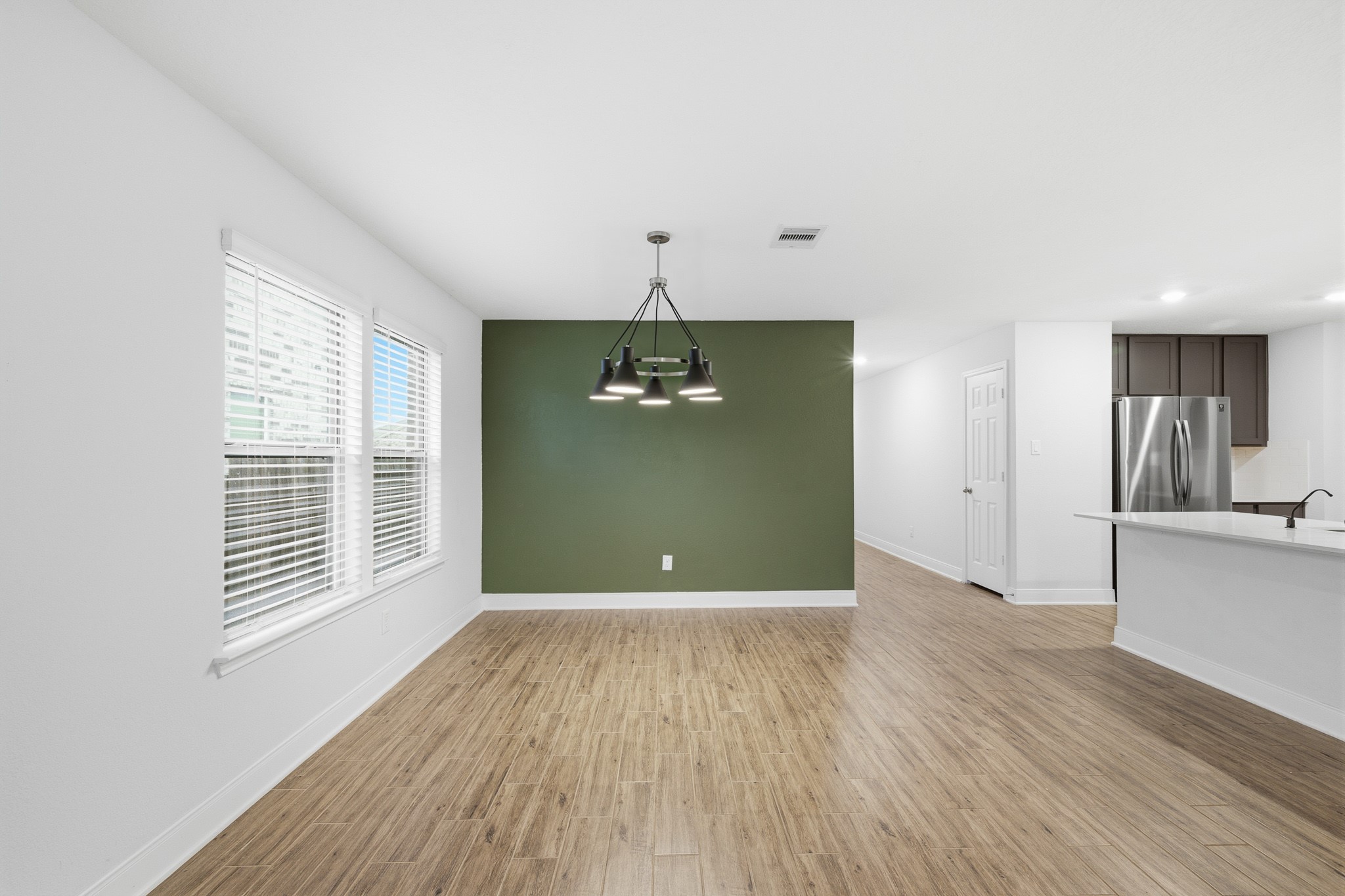 Bright and inviting dining area with sleek flooring, accented by a stylish green feature wall and modern light fixture, adjacent to an open-concept kitchen.